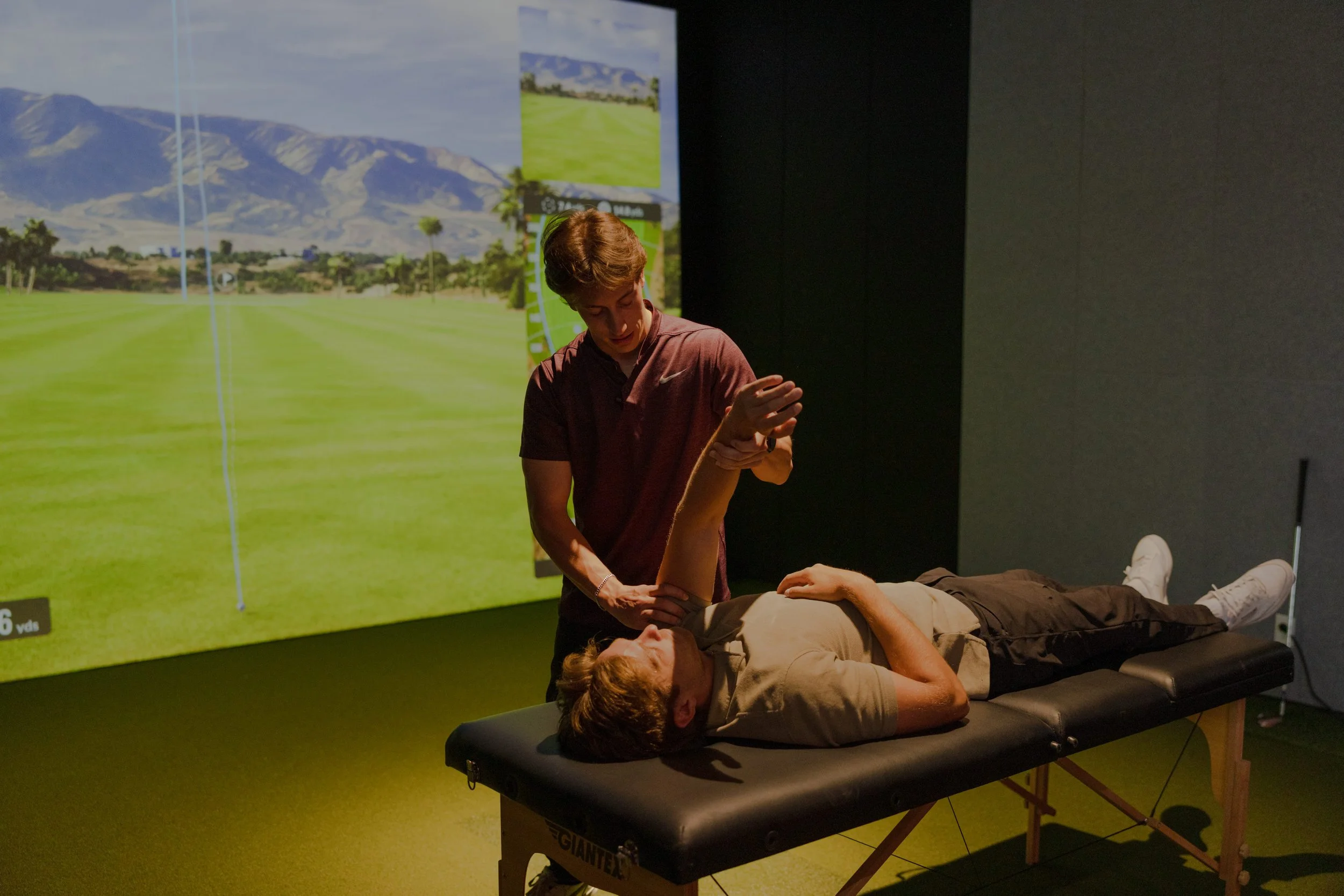 A practitioner assessing a golfers shoulder on a treatment table with a golf simulator screen behind