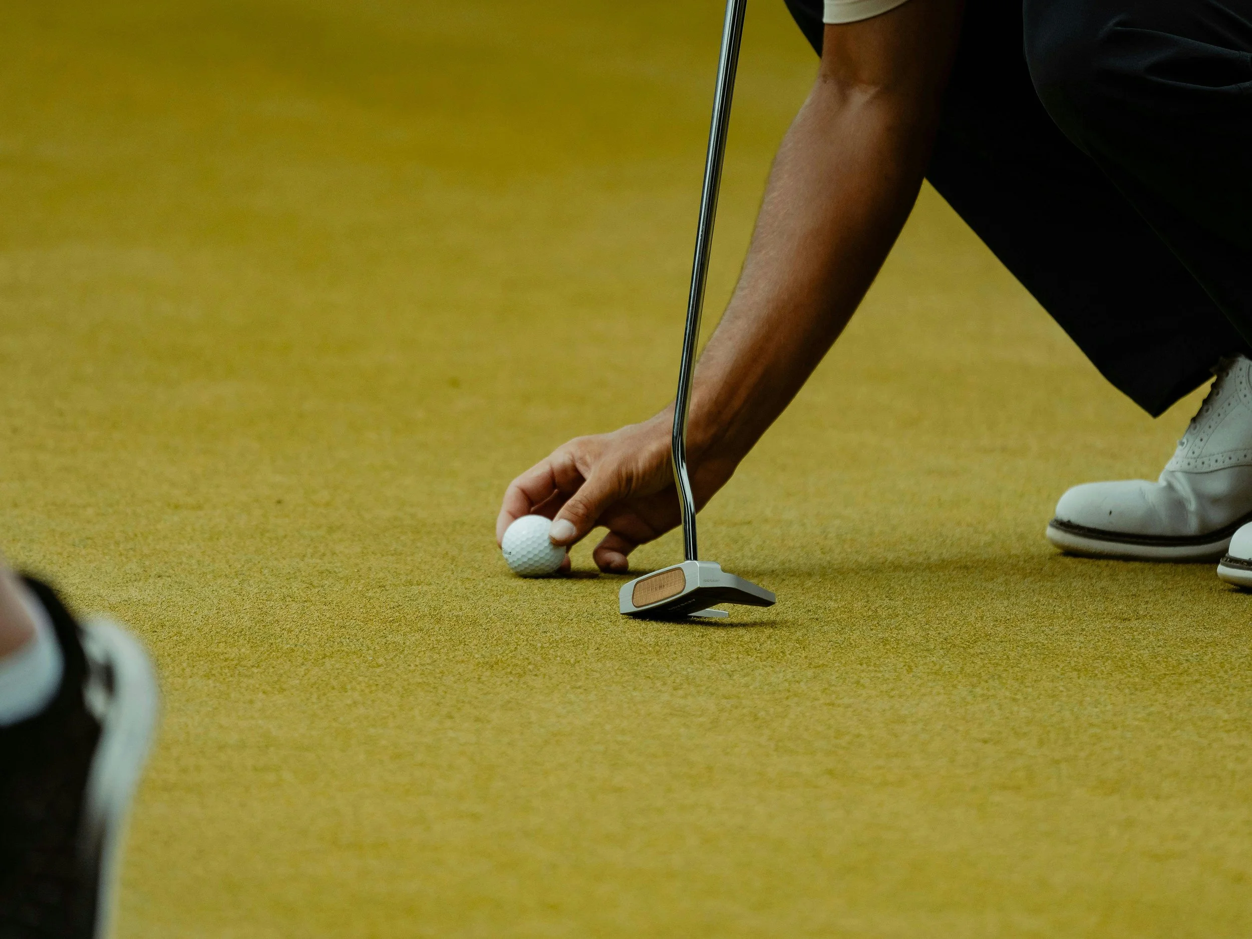 Person placing a golf ball on the ground while holding a putter, wearing white golf shoes and black clothing, on a yellow golf course or practice mat.