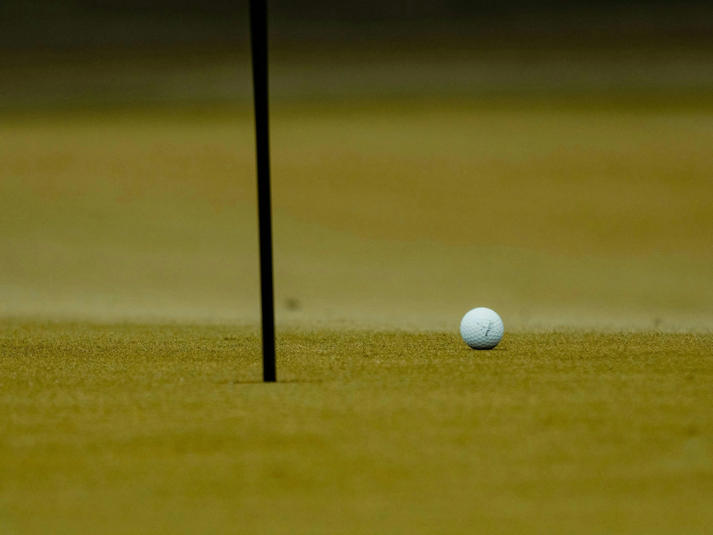 A golf ball on a putting green near a hole, with a flagstick marking the hole. The background is blurred.