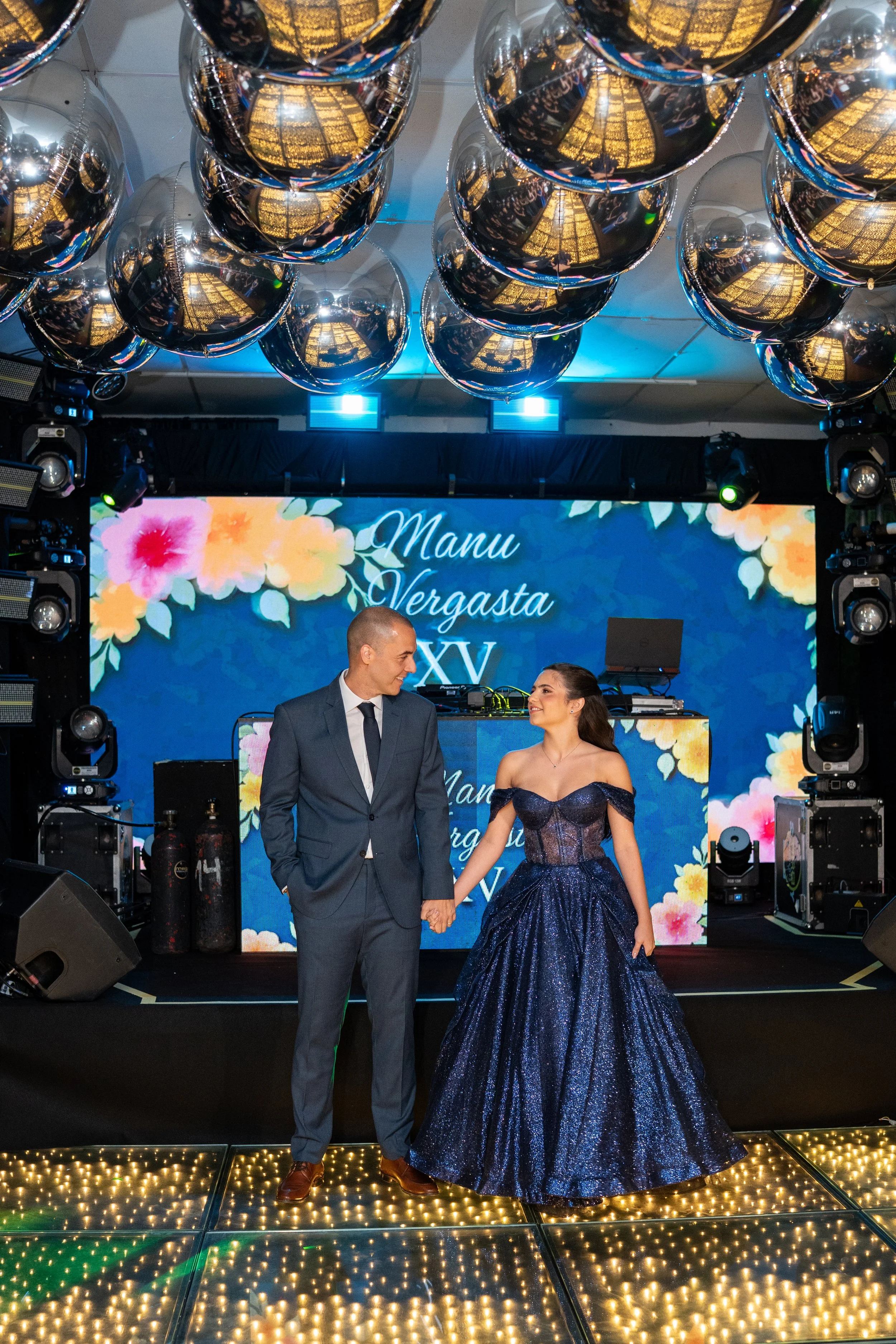 Casal segurando as mãos em um evento de comemoração, com um arranjo de balões metálicos dourados refletindo luz e uma tela de fundo com flores e o texto 'Manu Vergasta XV'.