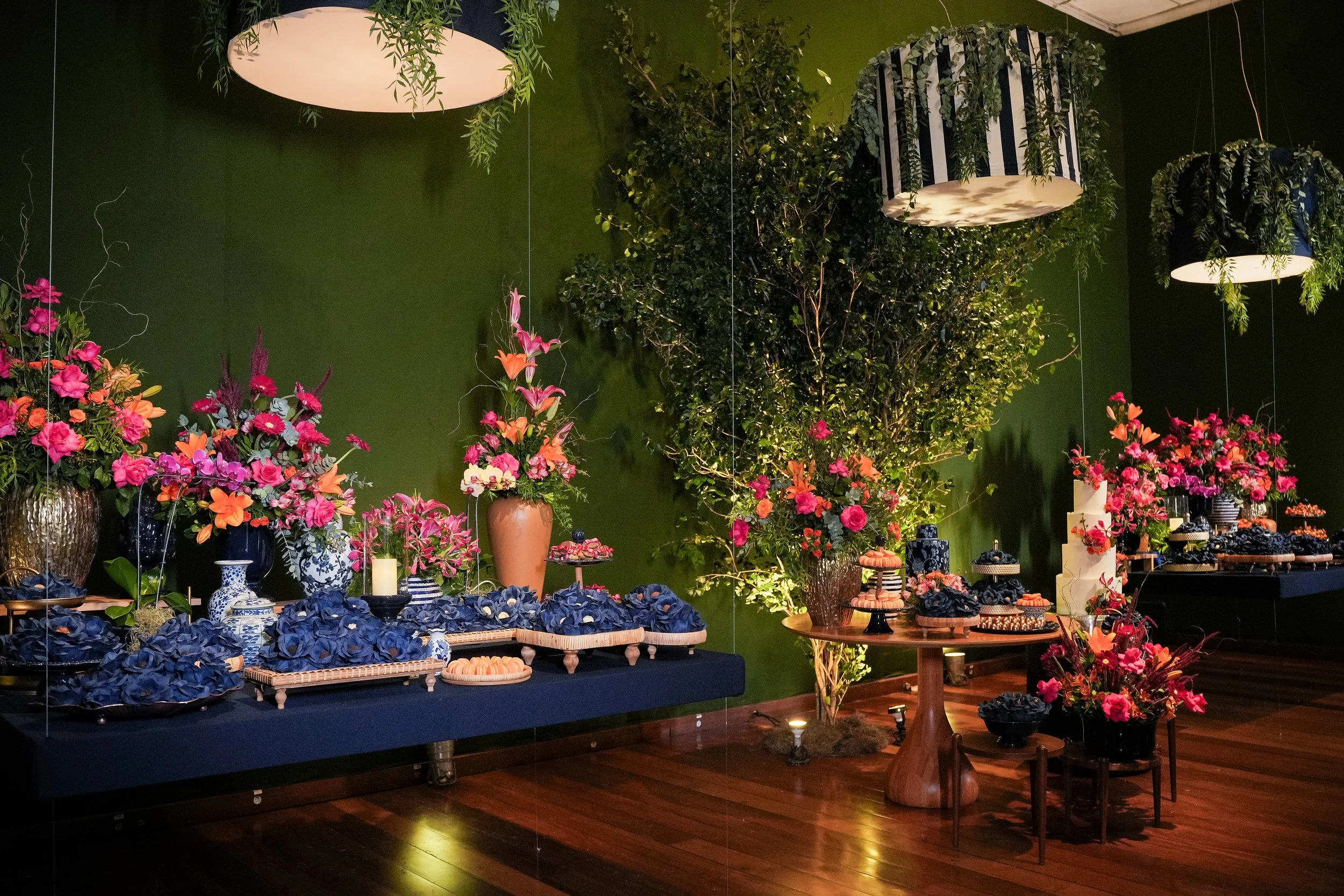 Decoração de festa com vasos de flores rosa, pratos de doces em cima de mesas e um fundo verde com plantas penduradas.