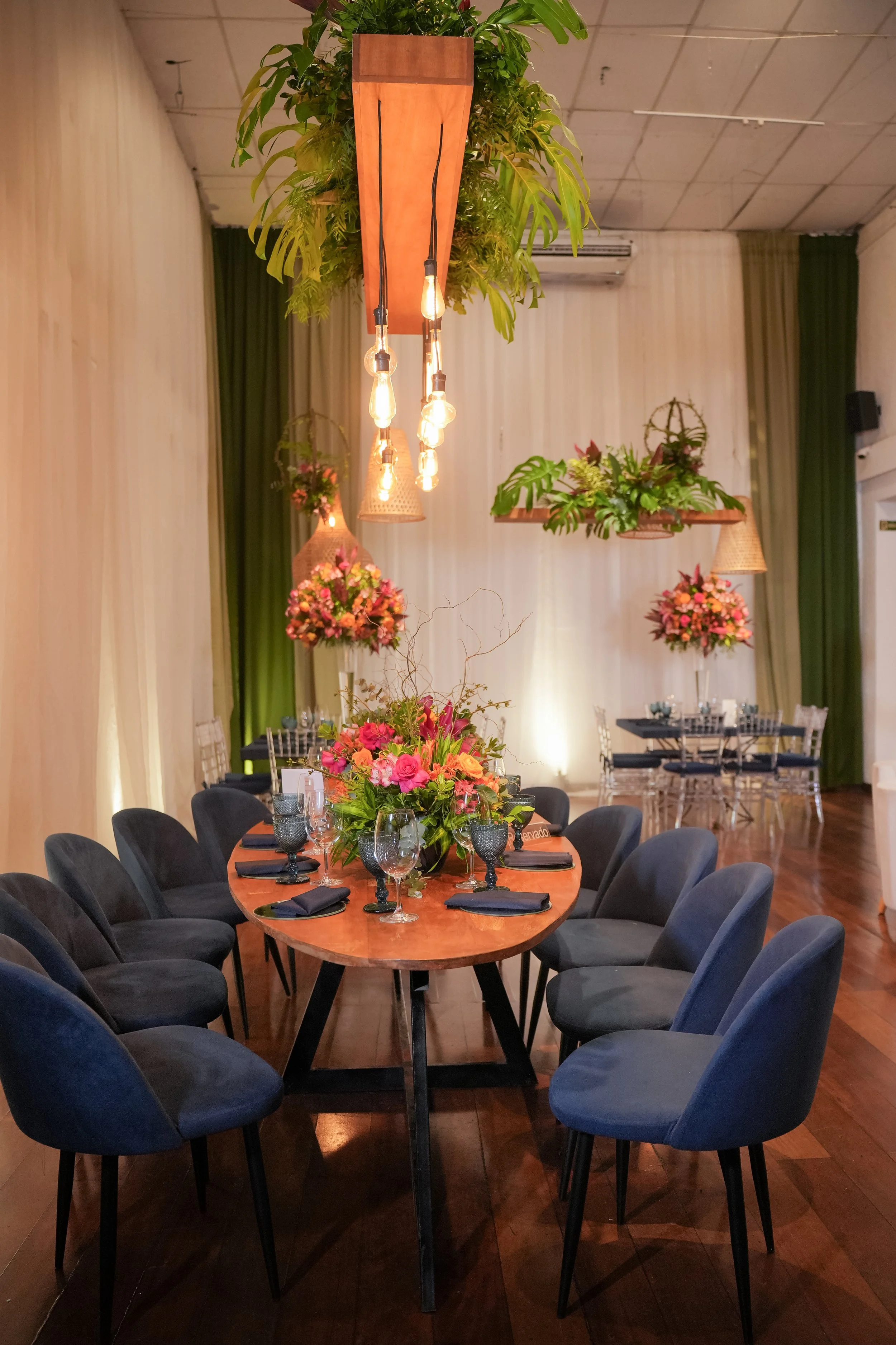 Sala de jantar decorada com mesa oval de madeira, cadeiras cinza, arranjos florais em tons de rosa e verde, iluminação suave, paredes brancas com cortinas verdes, e detalhes decorativos suspensos com plantas e luminárias.