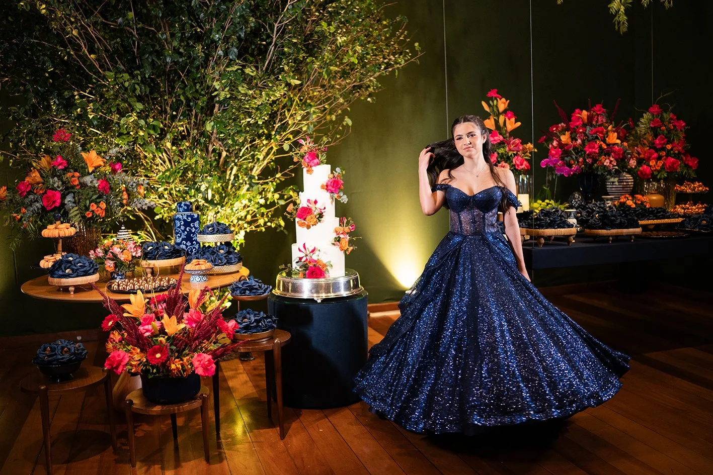 Juvenil em vestido azul brilhante, com decoração de festa com flores e bolo.
