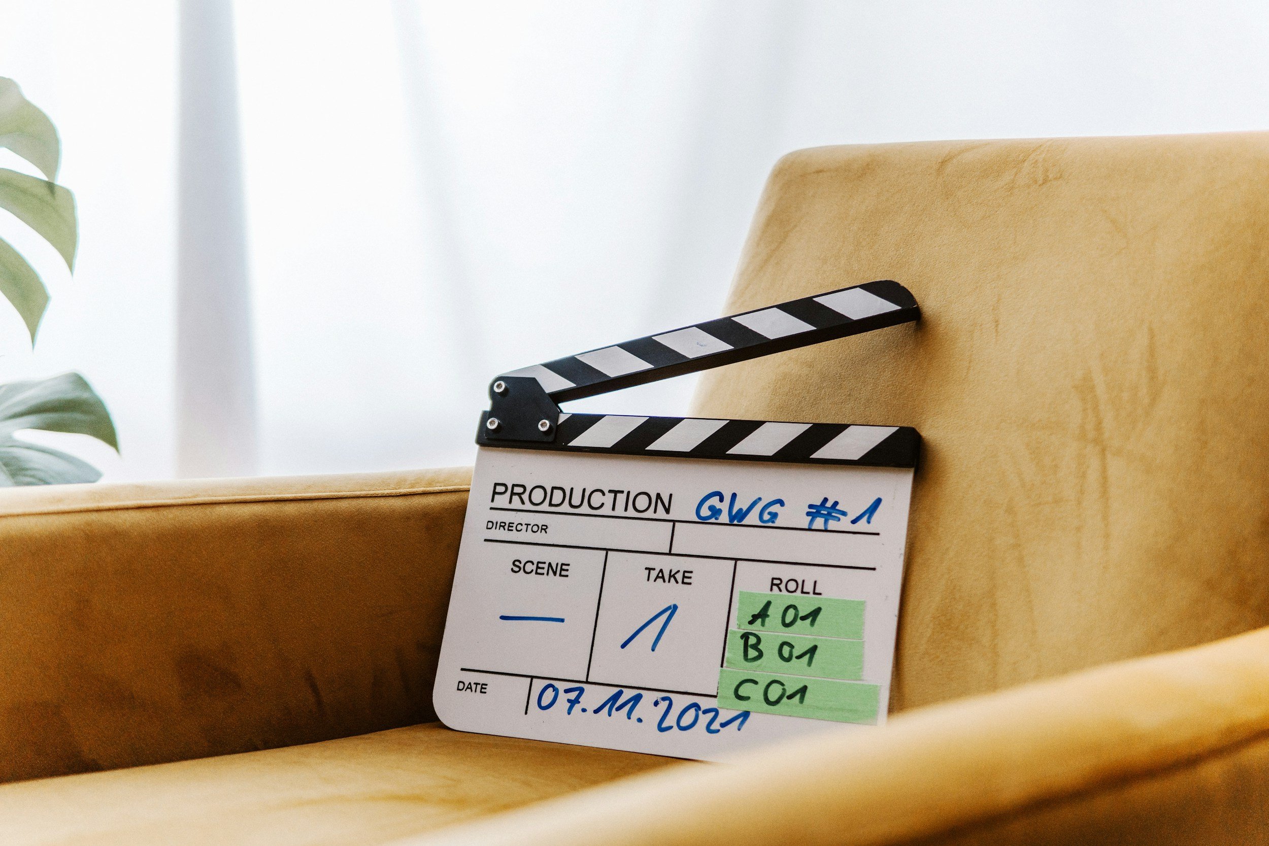 Clapperboard for production labeled "GWG #1" on a beige chair, with a plant partially visible on the left and a white background.