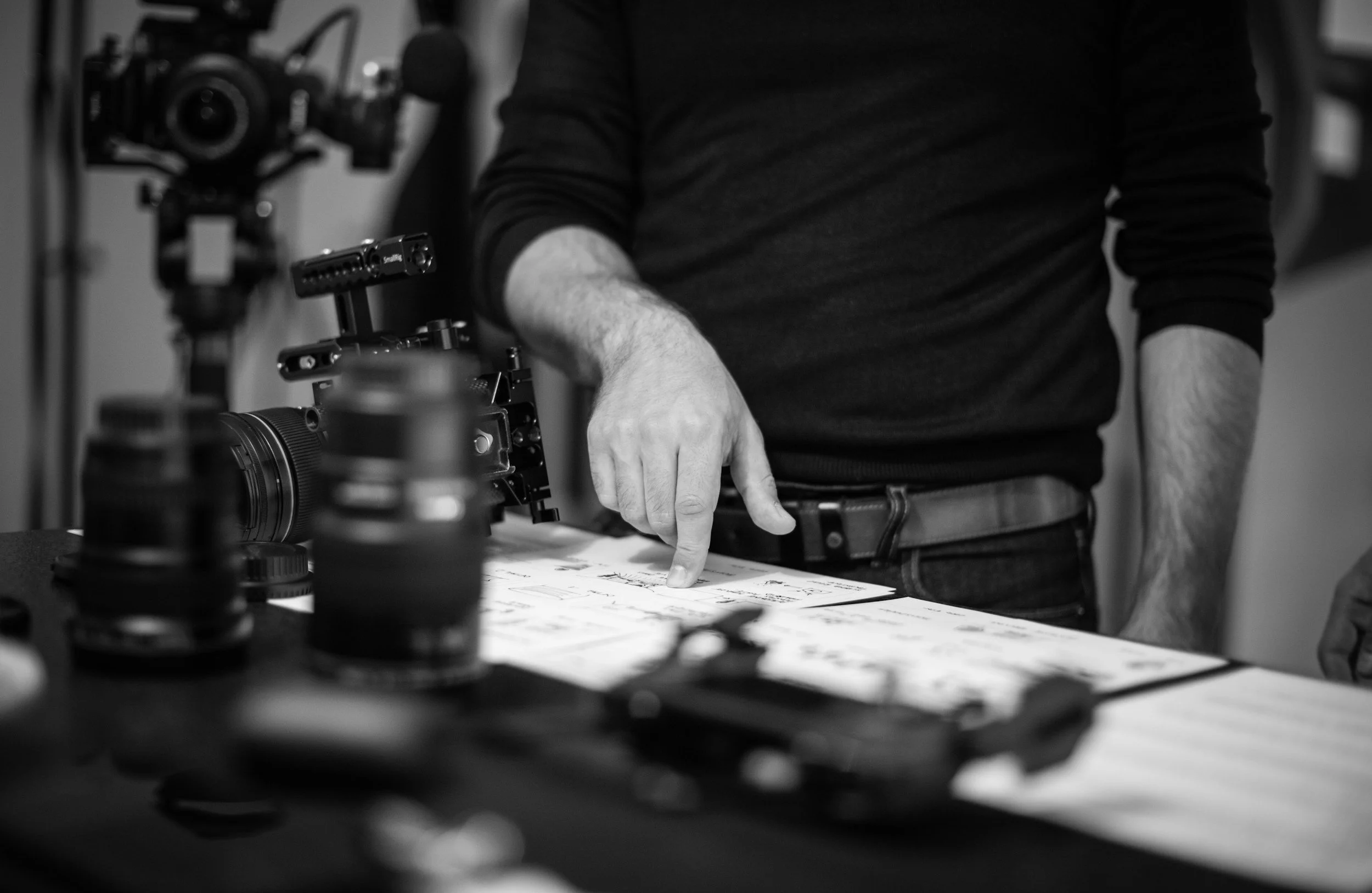 Black and white photo of a person wearing a dark long-sleeve shirt, pointing at a sheet of paper on a table, with camera equipment on the table and in the background.