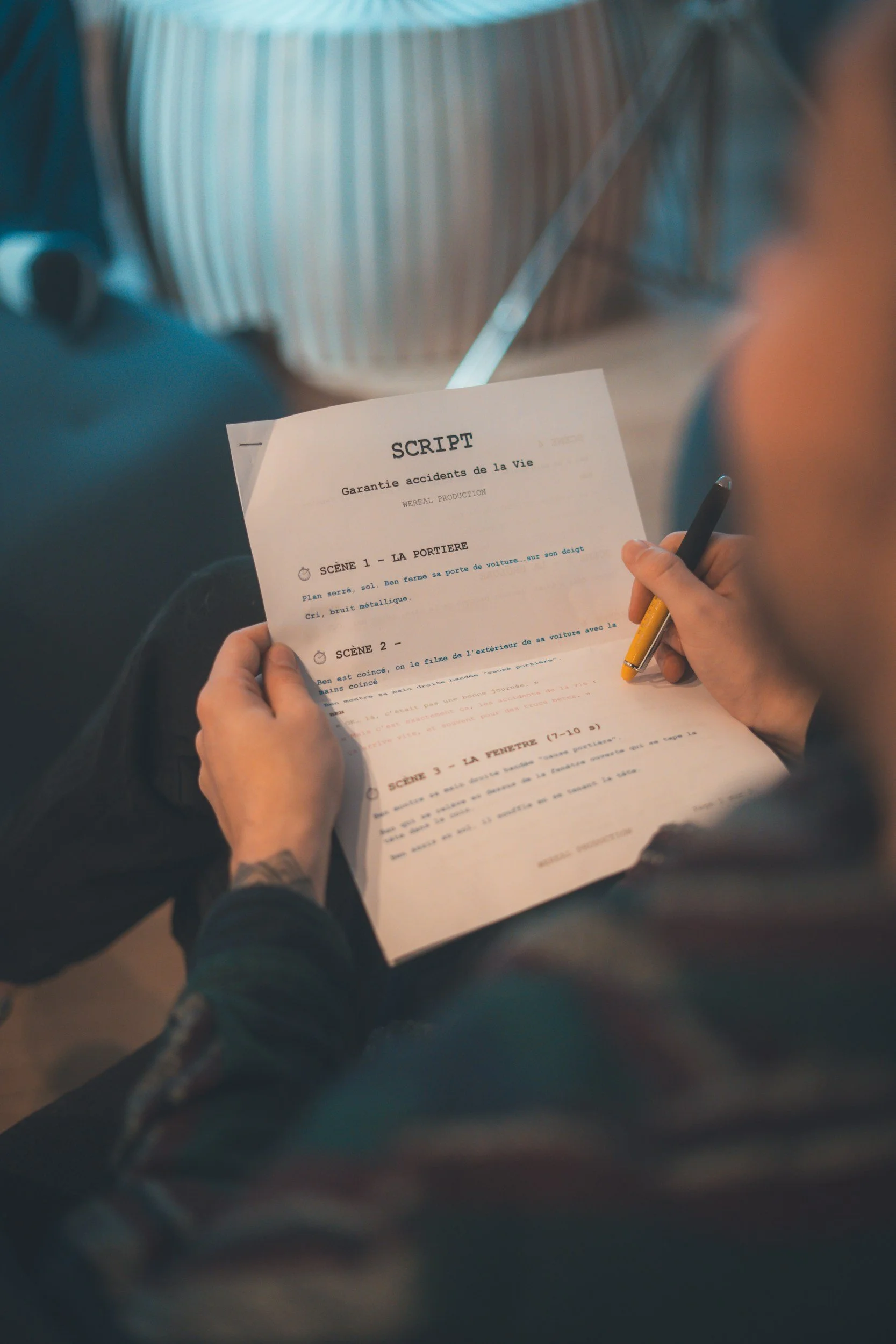 A person holding a script sheet with the title 'GARANTIE ACCIDENTS DE LA VIE' and working on a production alongside a person in the background. The person's hand is visible with a yellow and black pen.