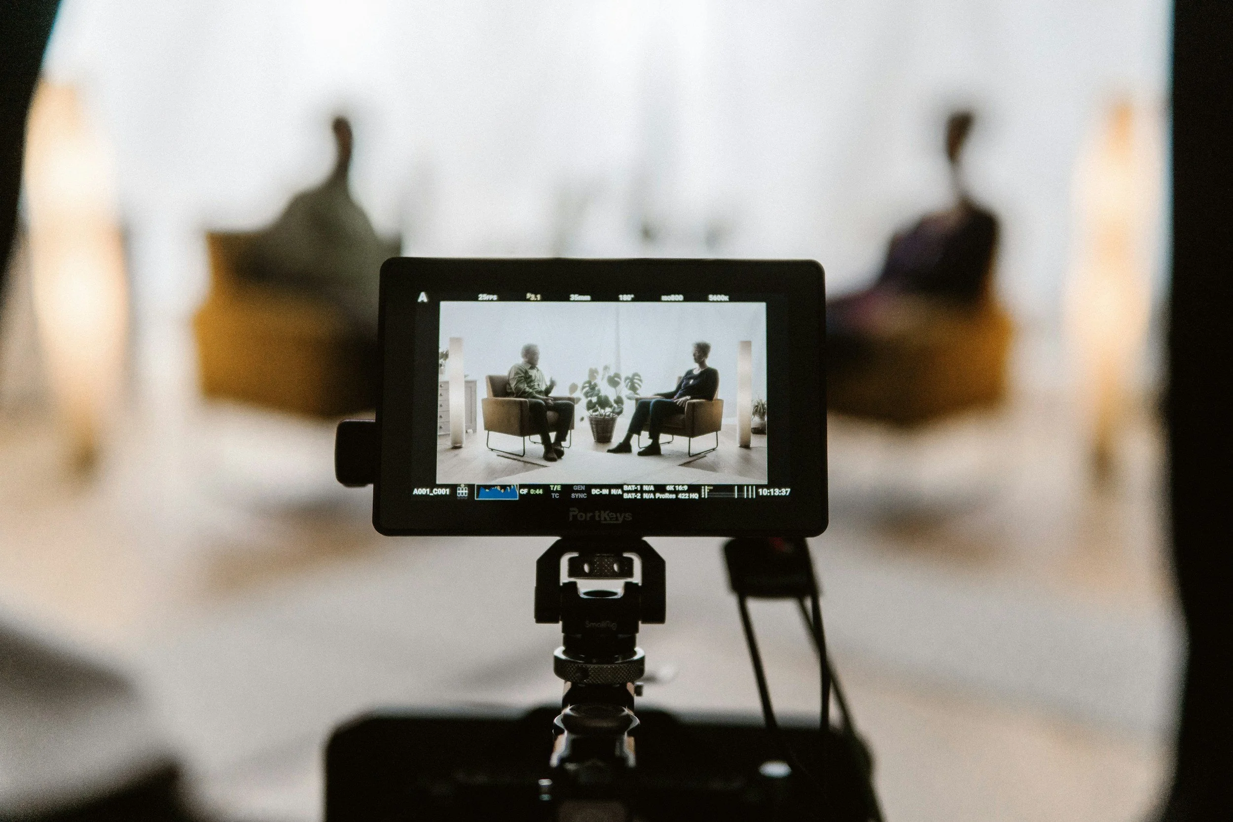 Photograph of a camera monitor showing two people seated in chairs, engaged in conversation, with a potted plant between them. The image is taken from behind the camera viewfinder.