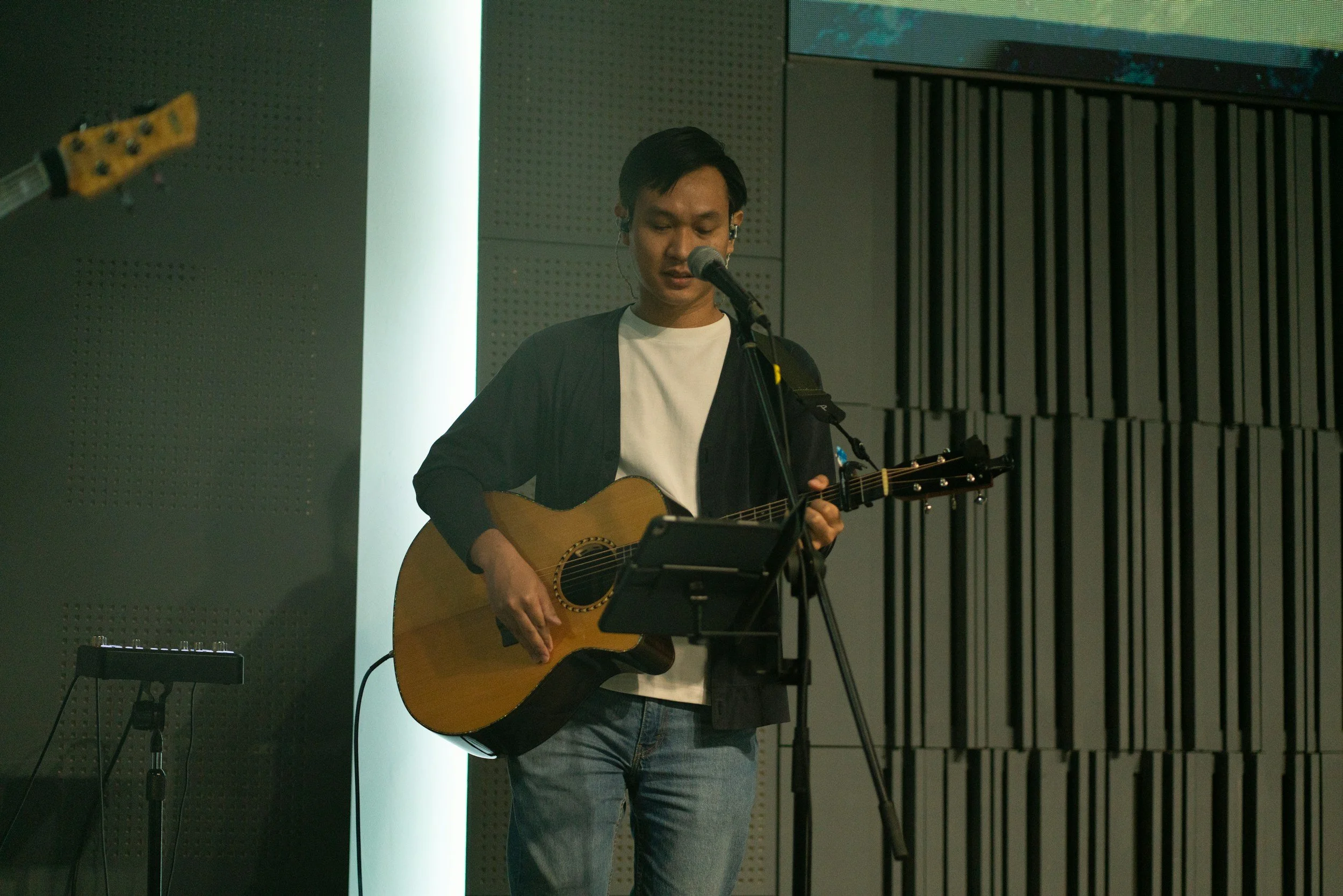 A young man with dark hair is playing an acoustic guitar and singing into a microphone during a performance in a modern indoor setting.