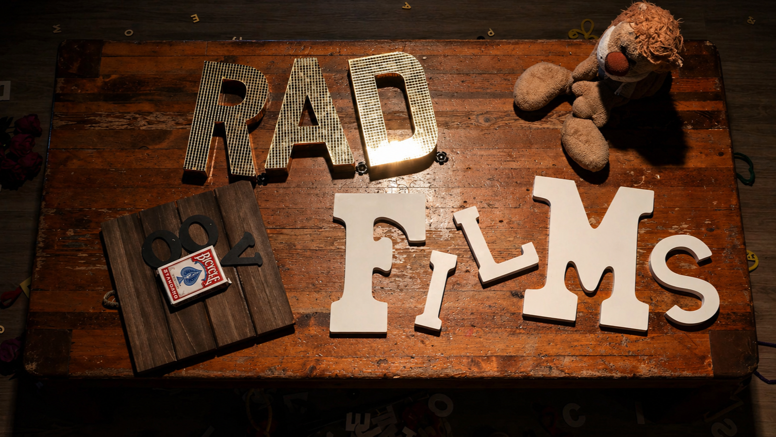 Wooden table with large decorative letters spelling 'DAD' with a light behind them and letters spelling 'FILS'. A teddy bear with a clown face and a box of playing cards are also on the table.
