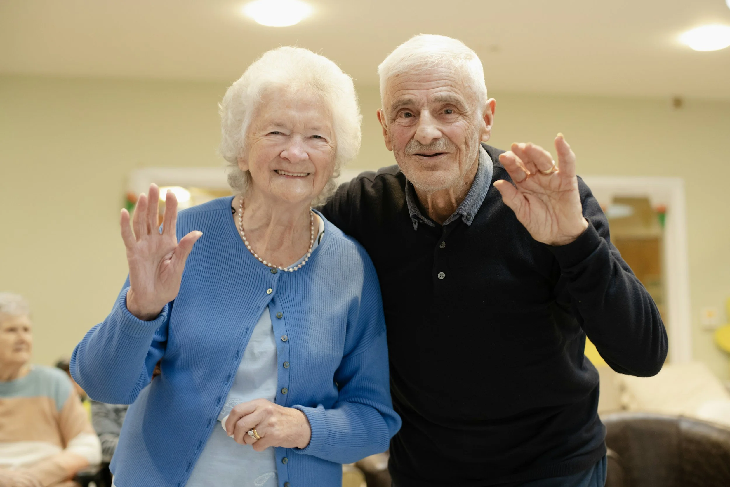 An elderly woman and a man smiling and waving at an indoor social gathering. The woman is wearing a blue sweater, pearl necklace, and has white hair. The man is wearing a dark sweater, has white hair, and a gray beard. They are standing close together, appearing friendly and cheerful.