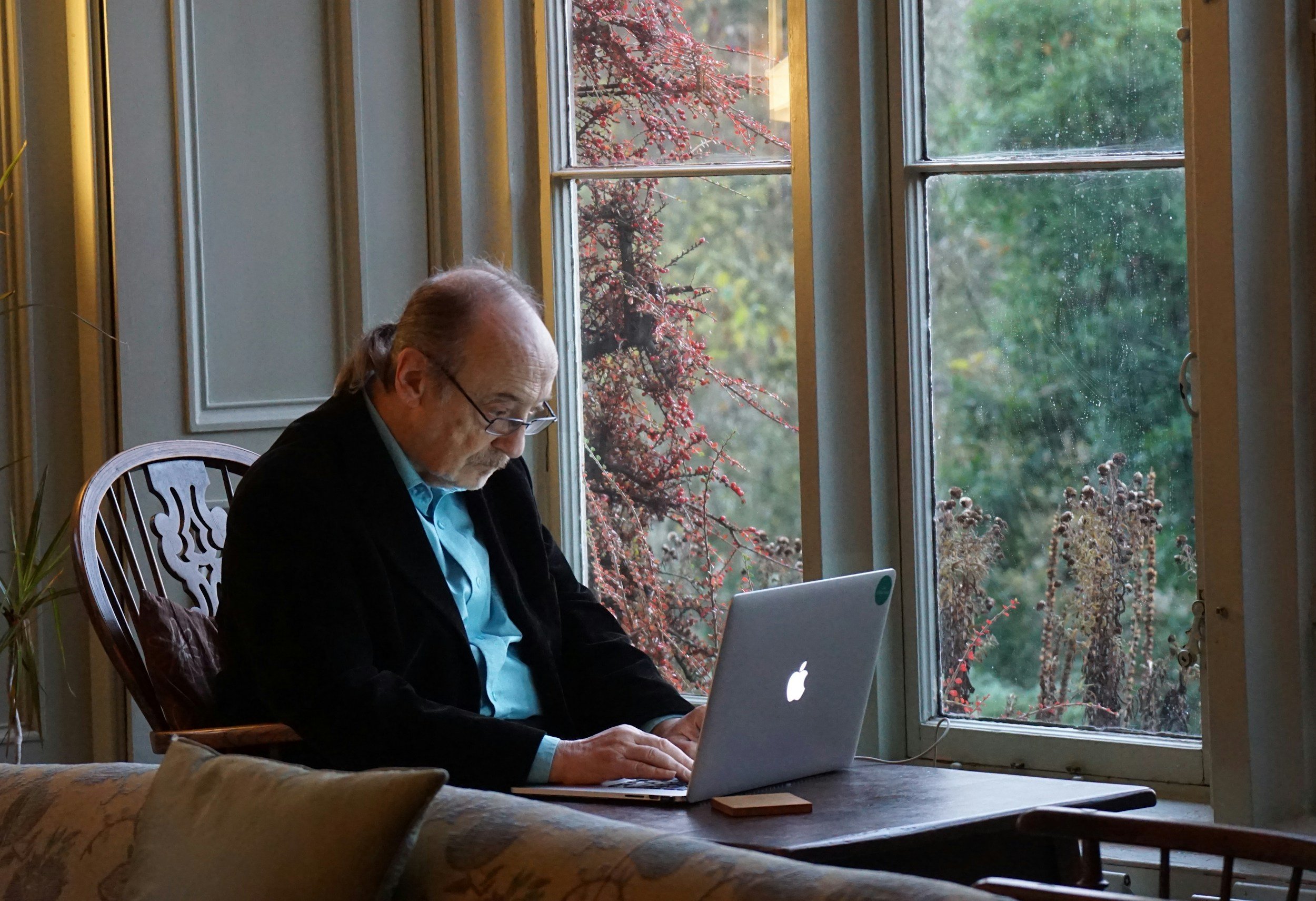An elderly man with glasses, a beard, and gray hair is sitting at a wooden table by a window with a view of trees and a flowering shrub outside. He is working on a silver laptop, dressed in a black blazer and blue shirt. Unspalsh image.