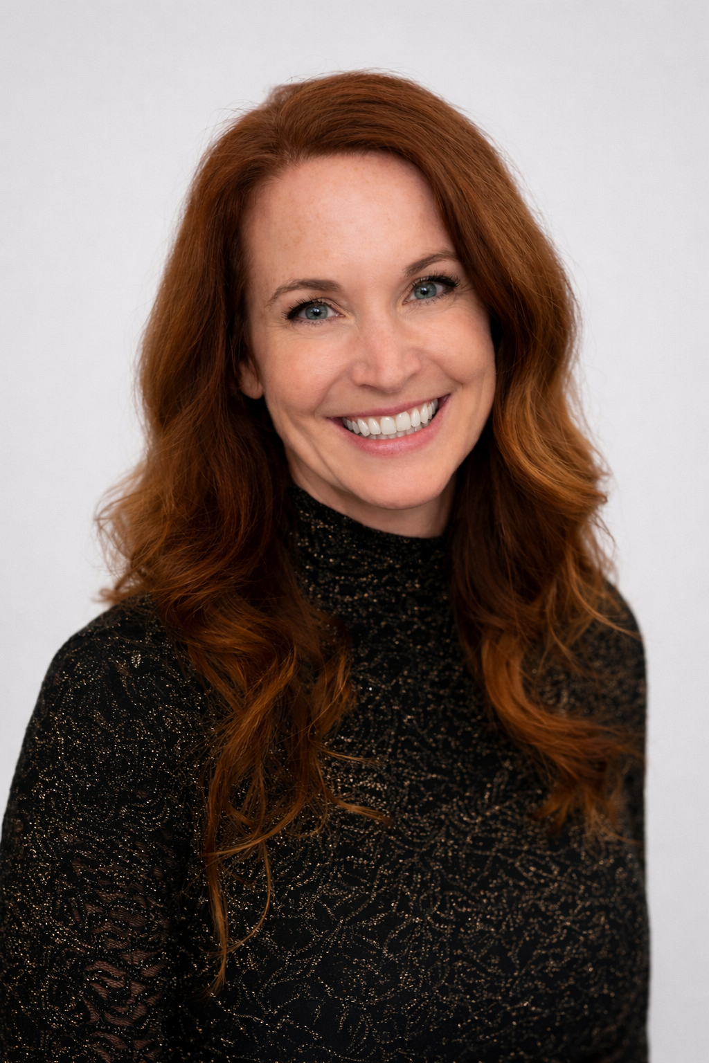Cori Headshot - Portrait of a smiling woman with red hair and blue eyes wearing a black, sparkly turtleneck against a plain white background.