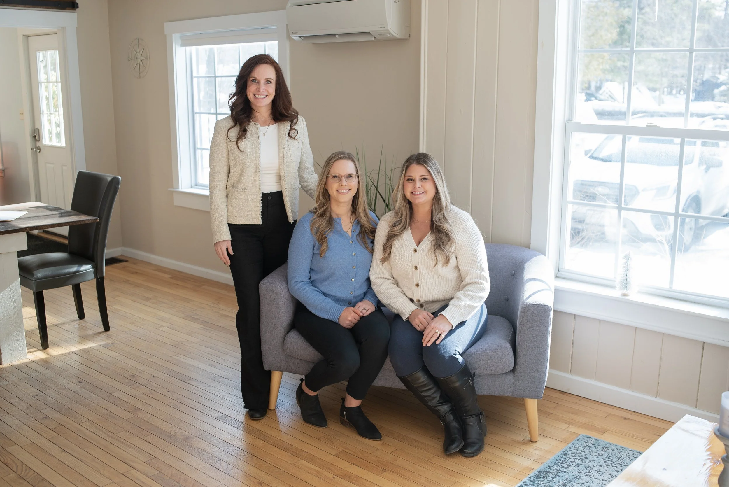 A woman standing and two women sitting on a gray couch in a bright room with large windows and wooden floors. Compass team image.