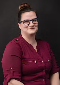 Katrina headshot - A woman with dark hair in a bun, wearing glasses and a burgundy shirt, smiling against a dark background.