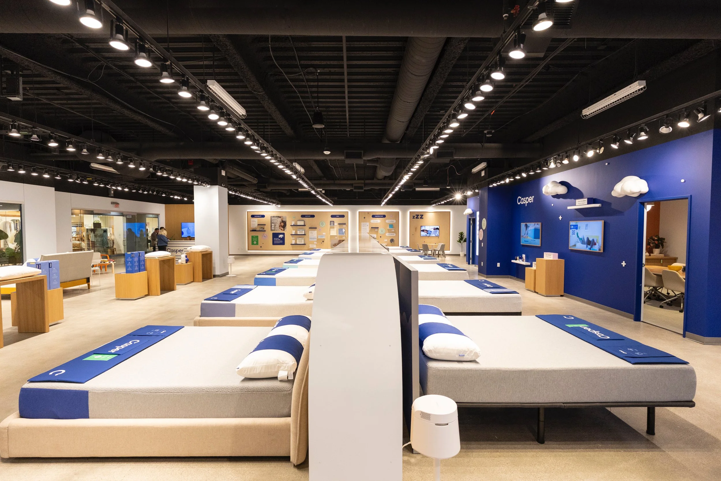 Showroom with beds, lounge chairs, and wall displays, under black ceiling with track lighting, blue and white color scheme.