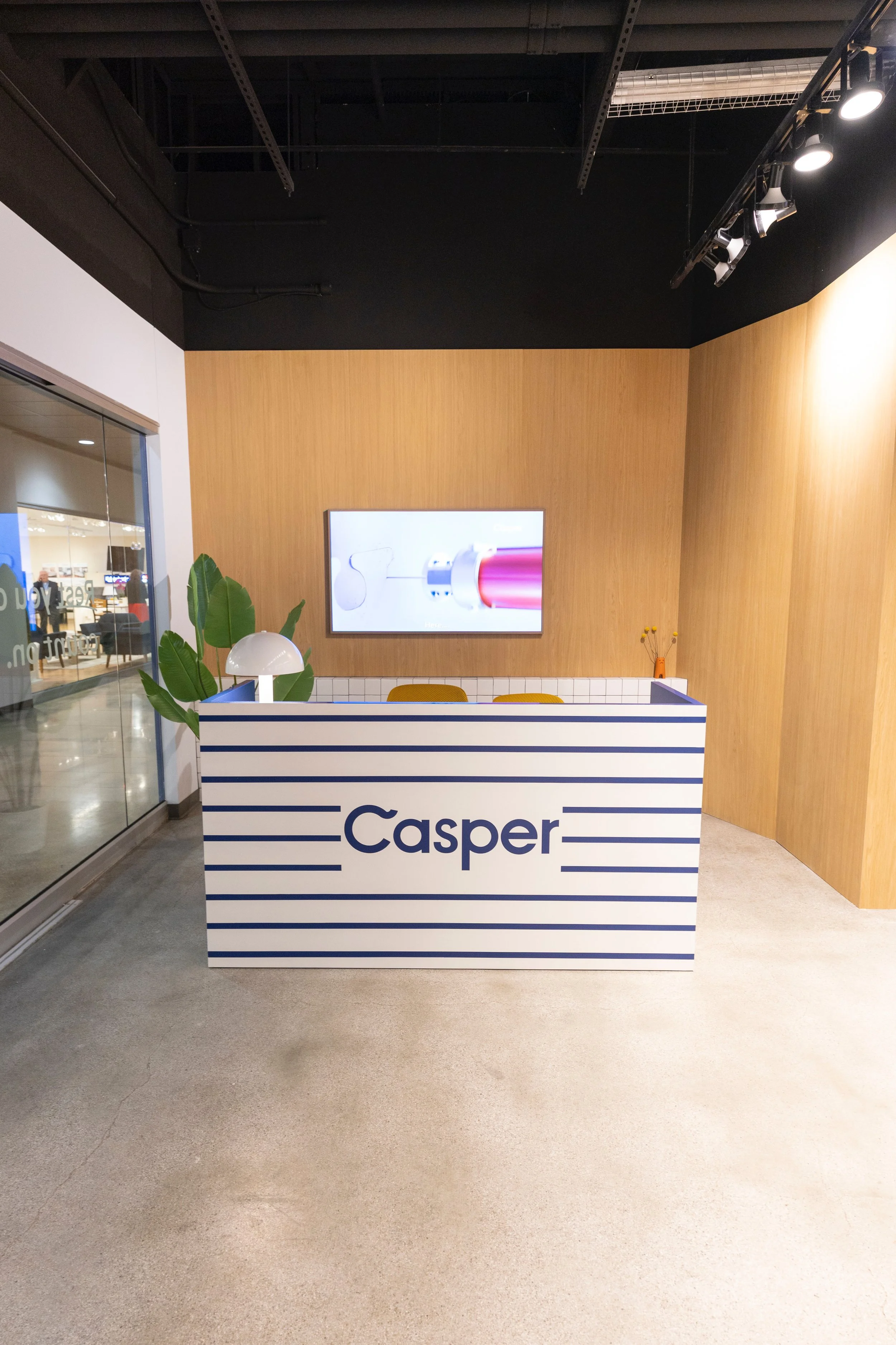A reception desk with the word 'Casper' in large letters, striped in white and blue, in a modern office space with wood wall panels, a wall-mounted screen, potted plant, and overhead lighting.