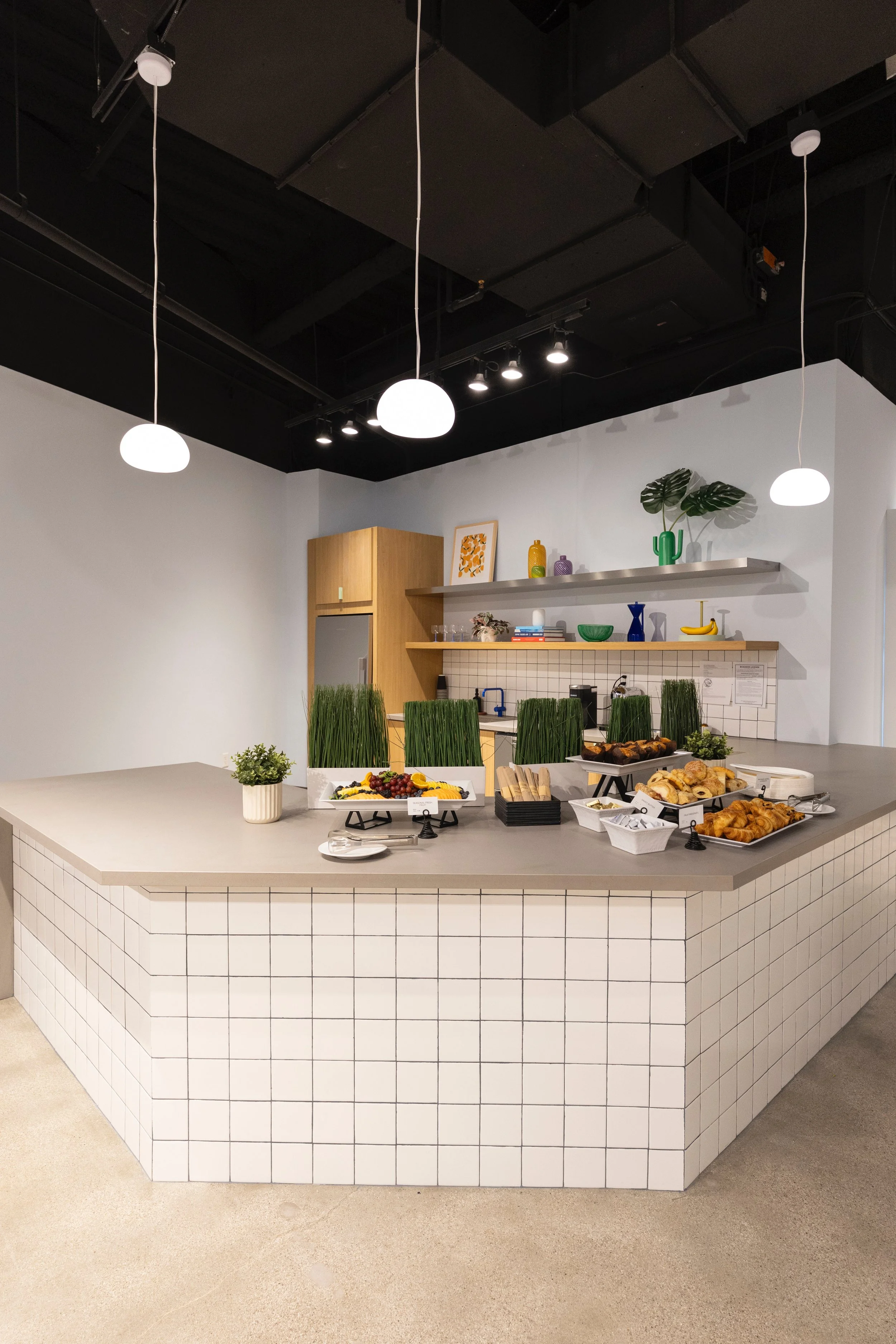 A modern kitchen counter with white tiled sides and a gray countertop, displaying various food items including pastries, fruit, and small plant arrangements. Behind the counter is a white wall with floating shelves holding decorative objects and plan