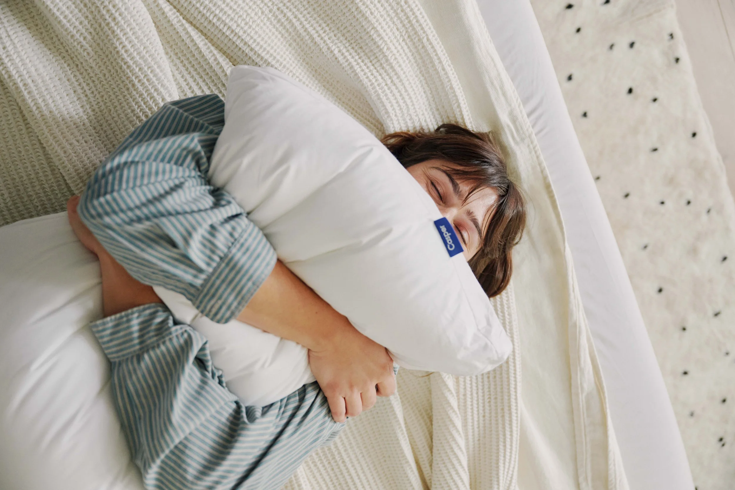 A person with brown hair and light skin is sleeping peacefully in bed, hugging a white pillow with a blue clothing tag, on a textured cream-colored blanket.