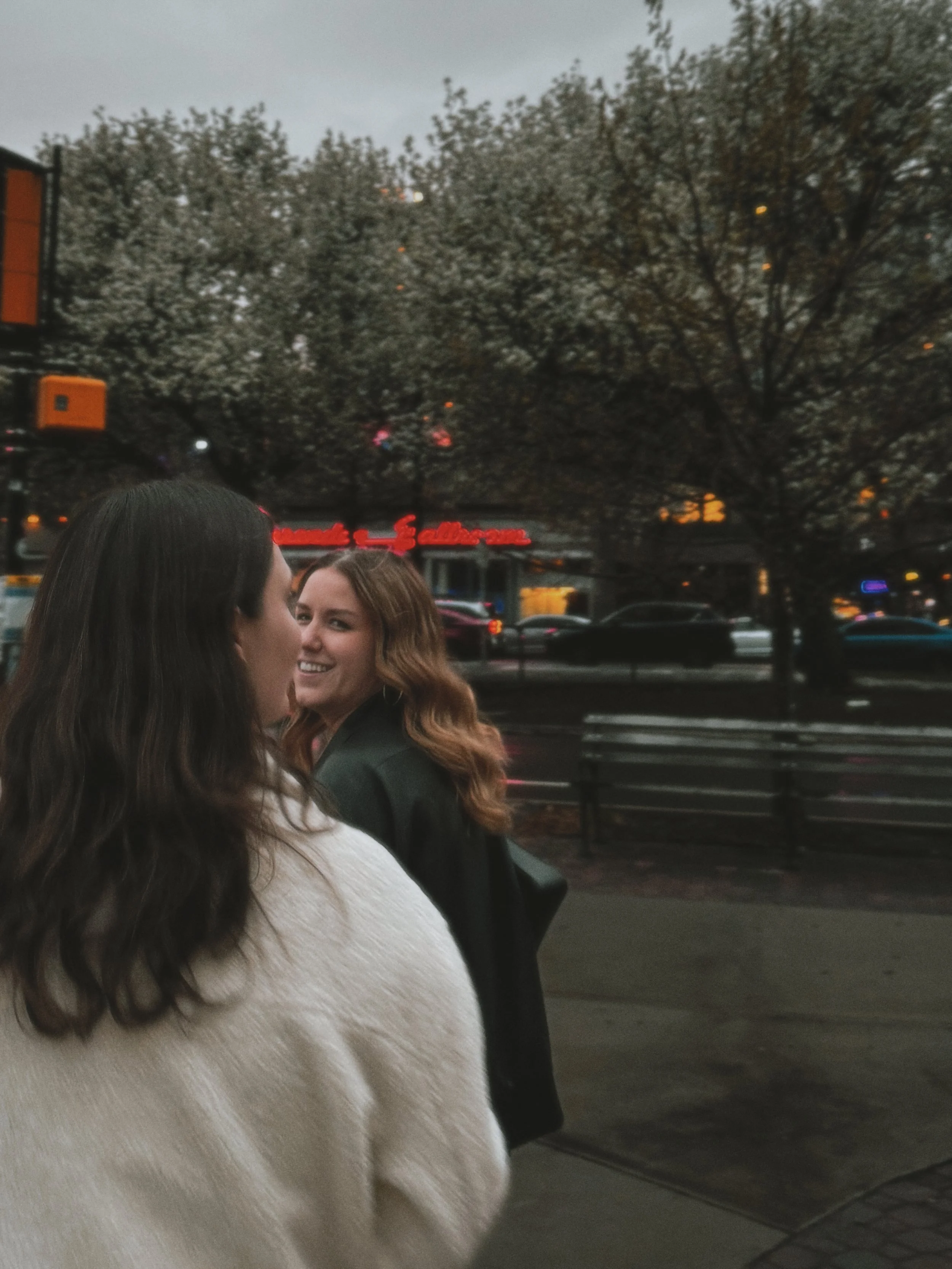 Two women standing outdoors near a busy street with cars, a tree with blossoms, and a building with illuminated signs in the background.