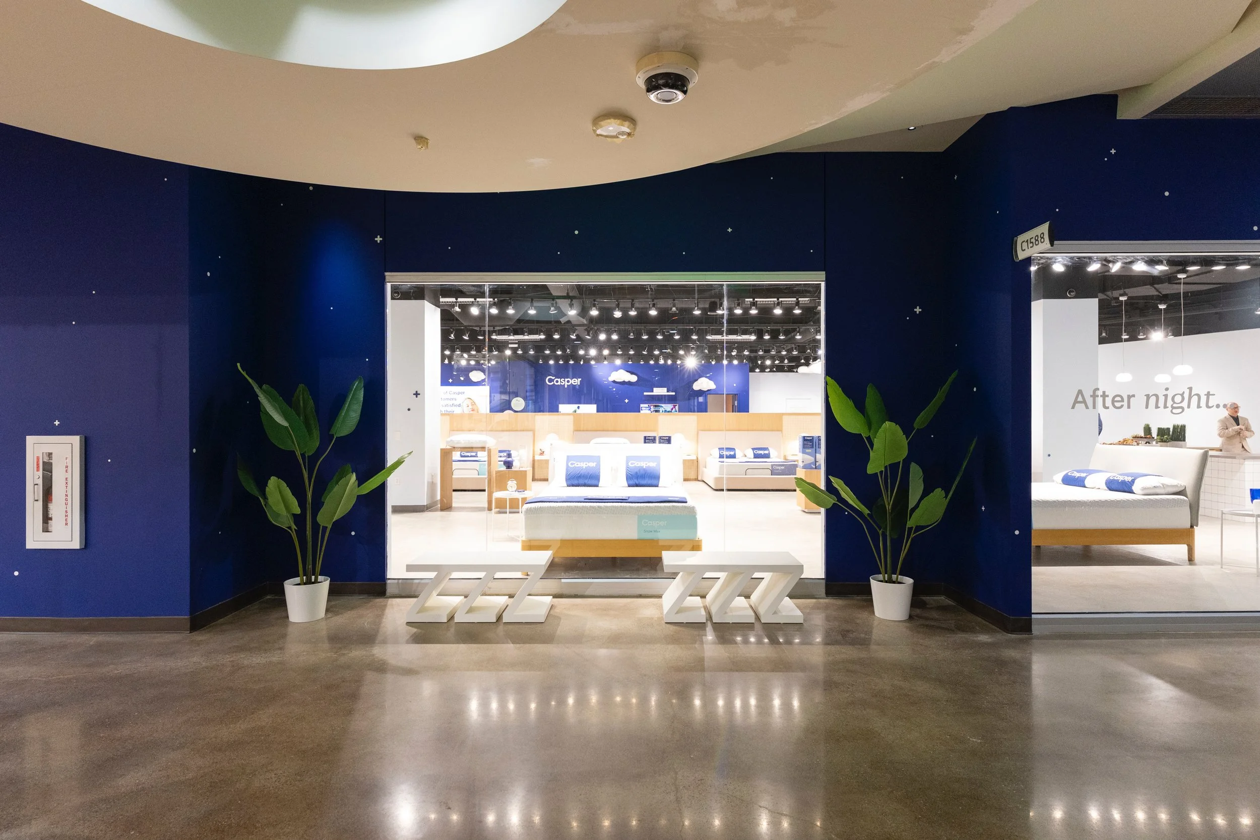 View of a furniture store display with a blue wall decorated with small scattered white stars, a glass entrance, and white furniture including beds and bedsheets. Two potted plants flank the entrance, and a person is standing on the right side near a white counter.