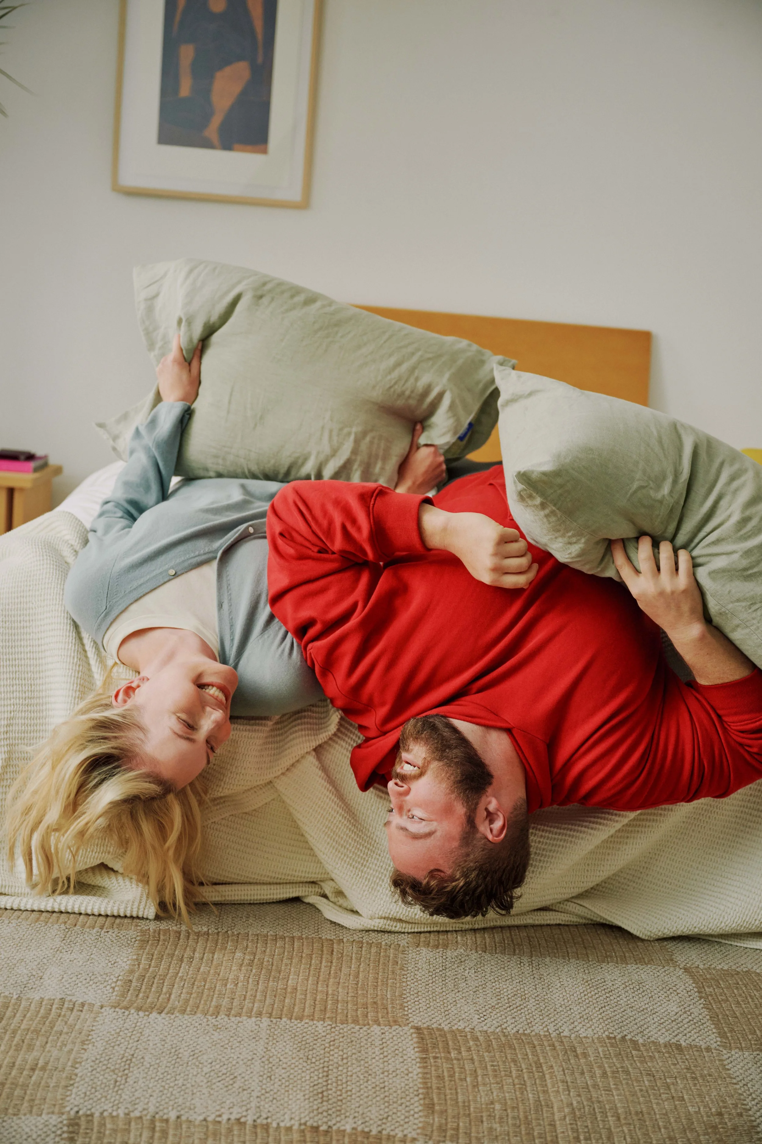 Four people are playfully wrestling on a beige sofa in a living room, smiling and laughing. One person is wearing a red shirt, another in gray, and two in light-colored tops. They are holding pillows and have a joyful expression.