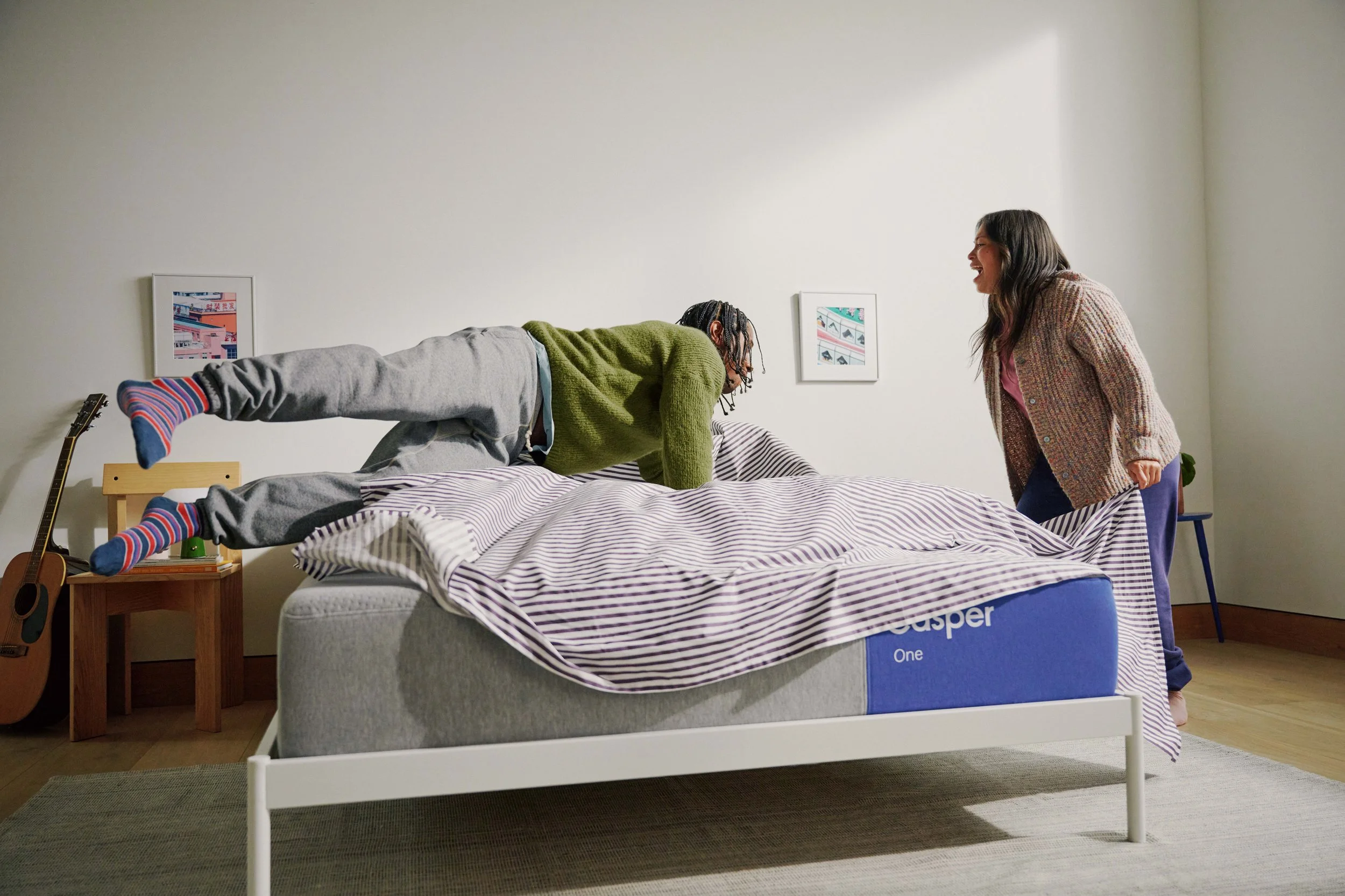 Two women laughing and playing on a bed in a bedroom. One woman is lying on her stomach on the bed, wearing a green sweater and gray sweatpants, while the other woman is standing and leaning towards her. The room has a minimalistic design with framed pictures on the white wall, a guitar leaning against the wall, and a blue box under the bed.