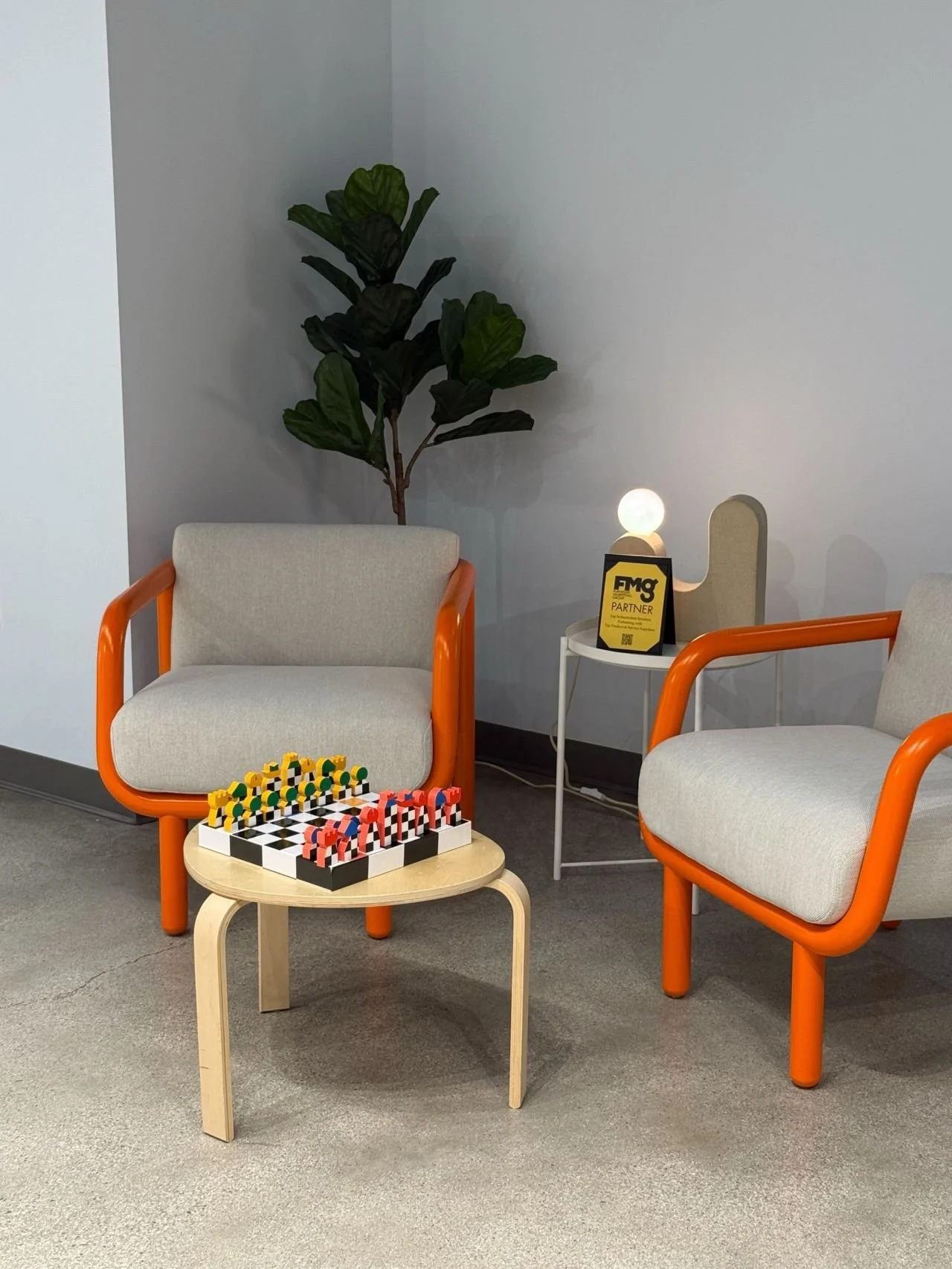 Two beige chairs with orange arms, a small wooden table with a chess set, a potted plant in the corner, and a white side table with a lamp and a yellow sign reading 'FMG Partner' in an indoor setting.