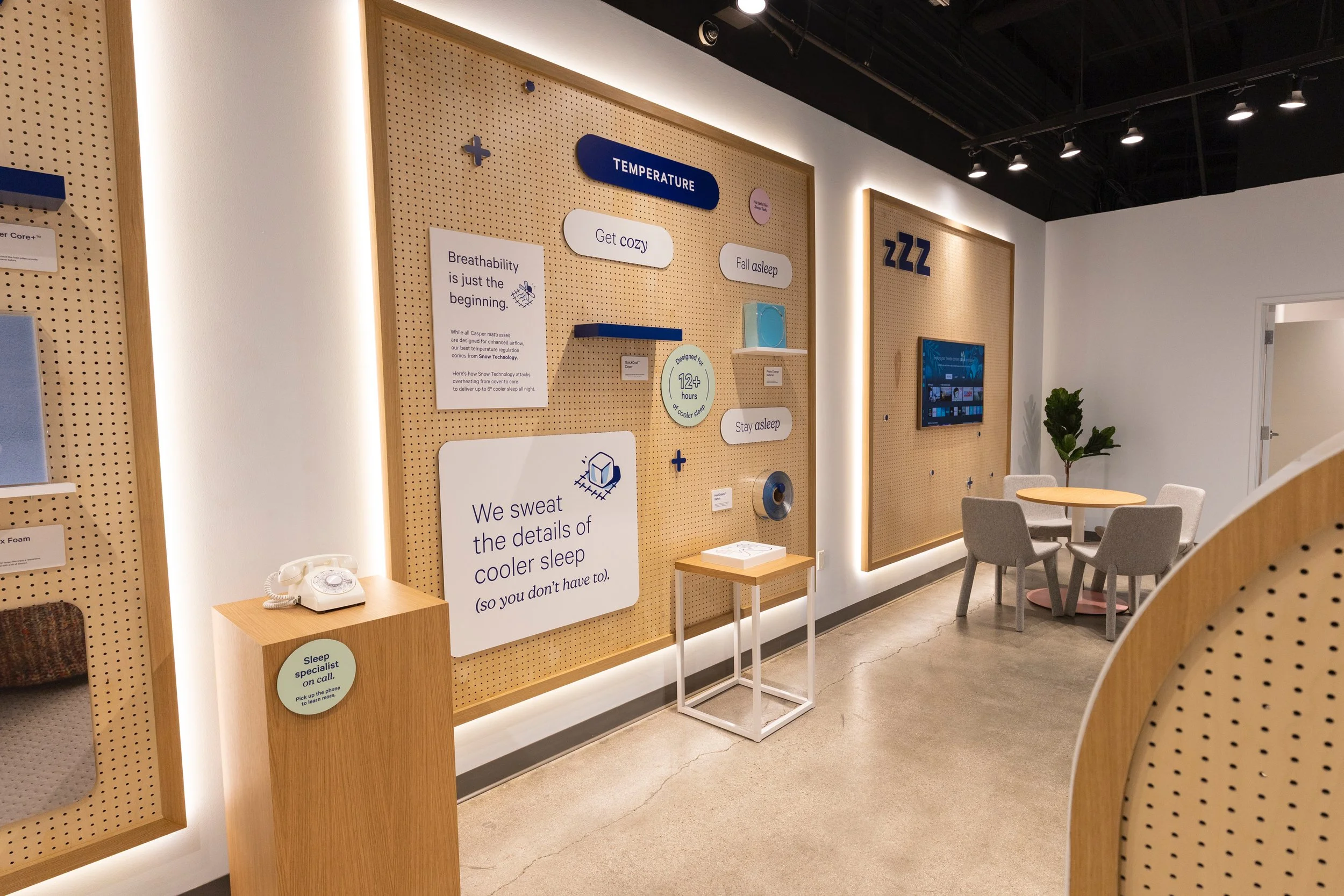 Interior of a store display area with signboards related to sleep and comfort, including a small round table, chairs, a potted plant, and informational posters about mattresses and sleep technology.