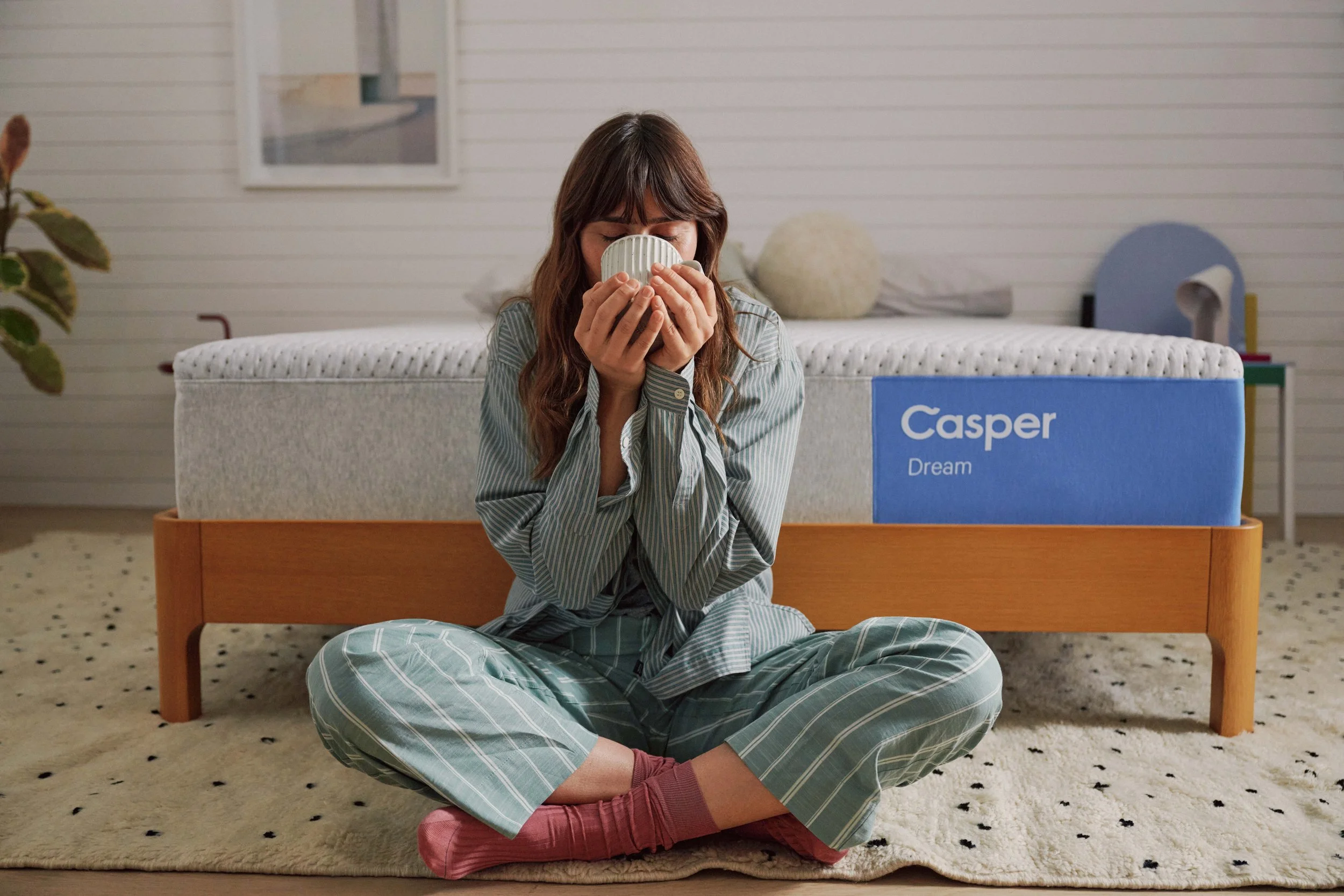 A woman sitting cross-legged on a patterned rug, holding a mug with both hands, dressed in matching striped pajamas, with a bed in the background that has a blue box labeled 'Casper Dream' and a fluffy pillow.