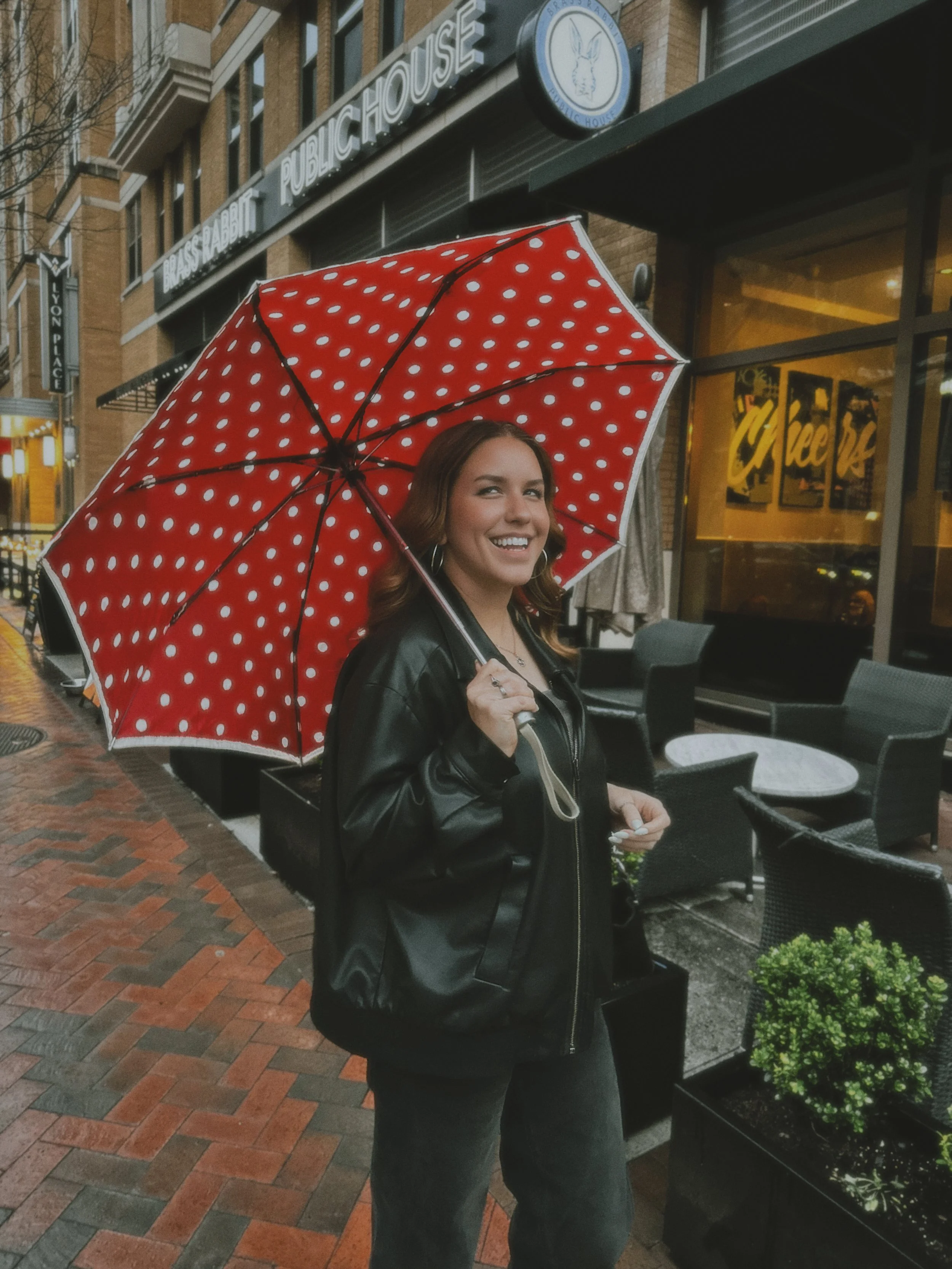 A smiling woman with brown hair holding a red umbrella with white polka dots stands on a brick sidewalk outside a restaurant. The restaurant has an outdoor seating area with black tables and chairs, and a large window displaying the word 'Cheers'. The building behind her has a sign that reads 'Blaise Pascal Public House' with a blue circular logo featuring a rabbit or hare outline.