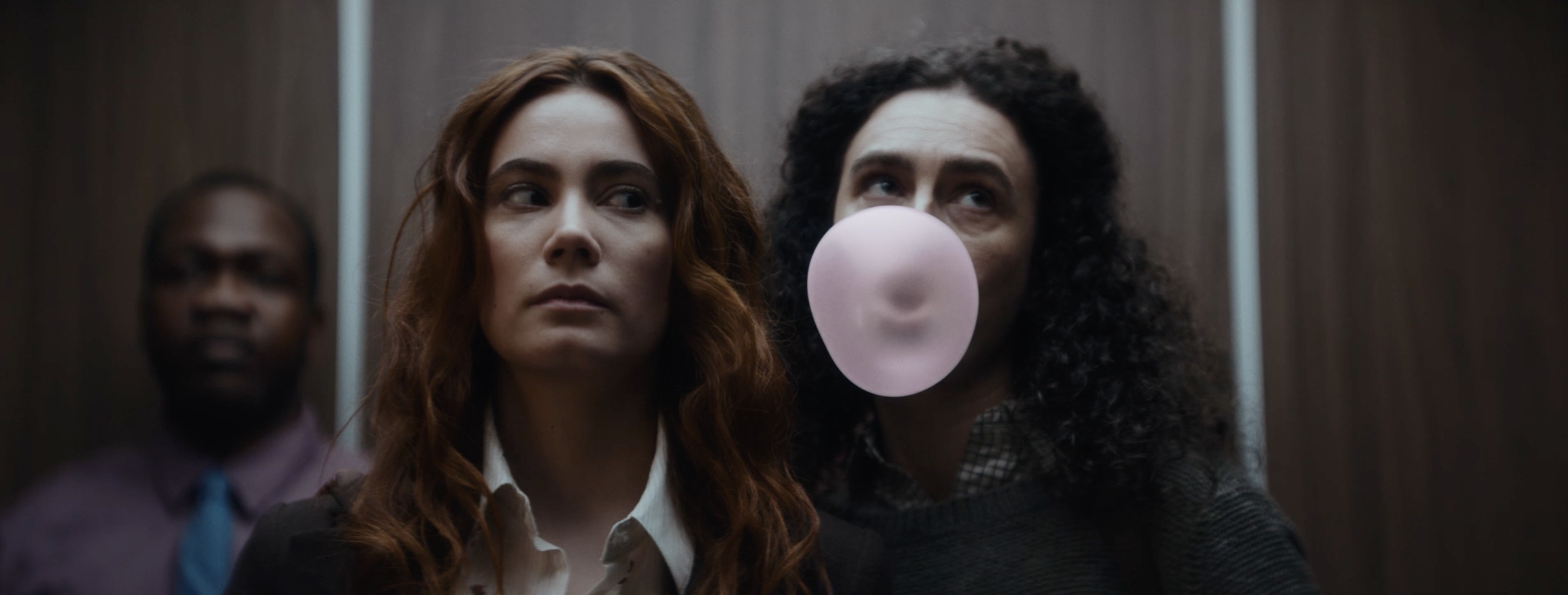 Three women standing in an elevator, two of them are in focus, one behind them is blurred. The woman on the right is blowing pink bubblegum.