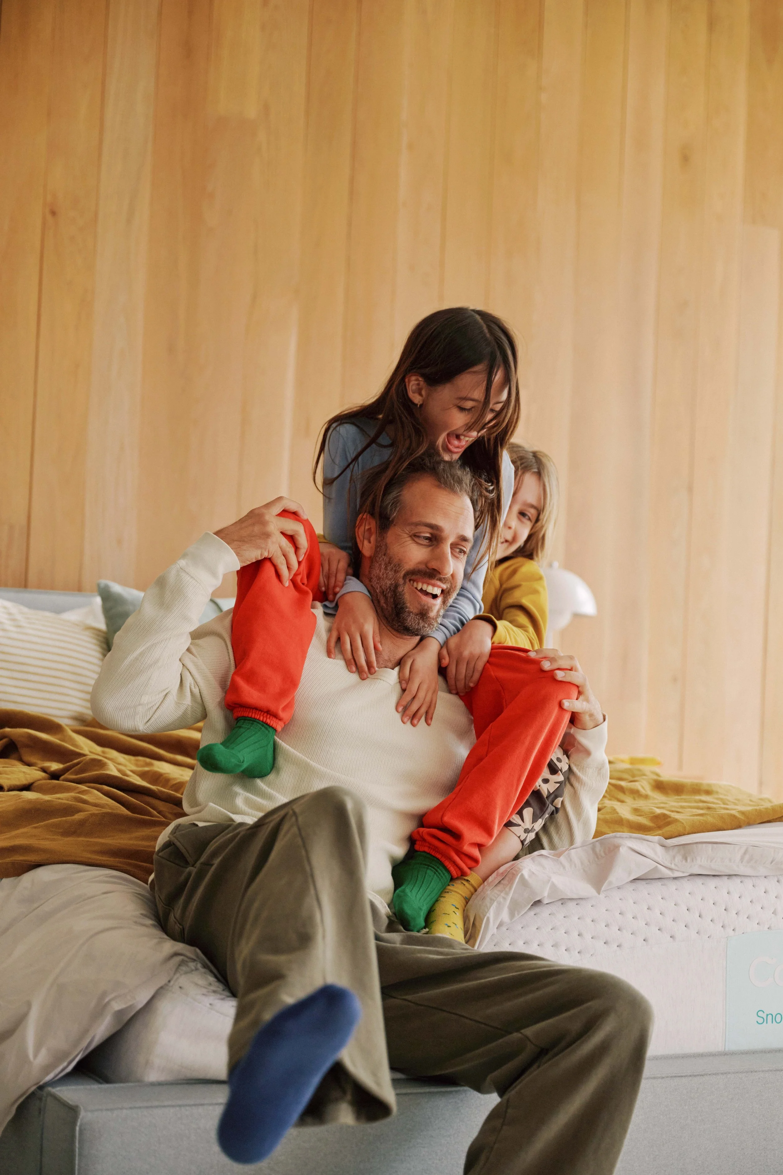 A man and two young girls sitting on a bed. The man is smiling while the girls are laughing and playfully crawling over him. One girl wears a blue shirt, and the other girl wears a yellow shirt. The scene is inside a bedroom with wooden walls, and there are pillows and a blanket on the bed.