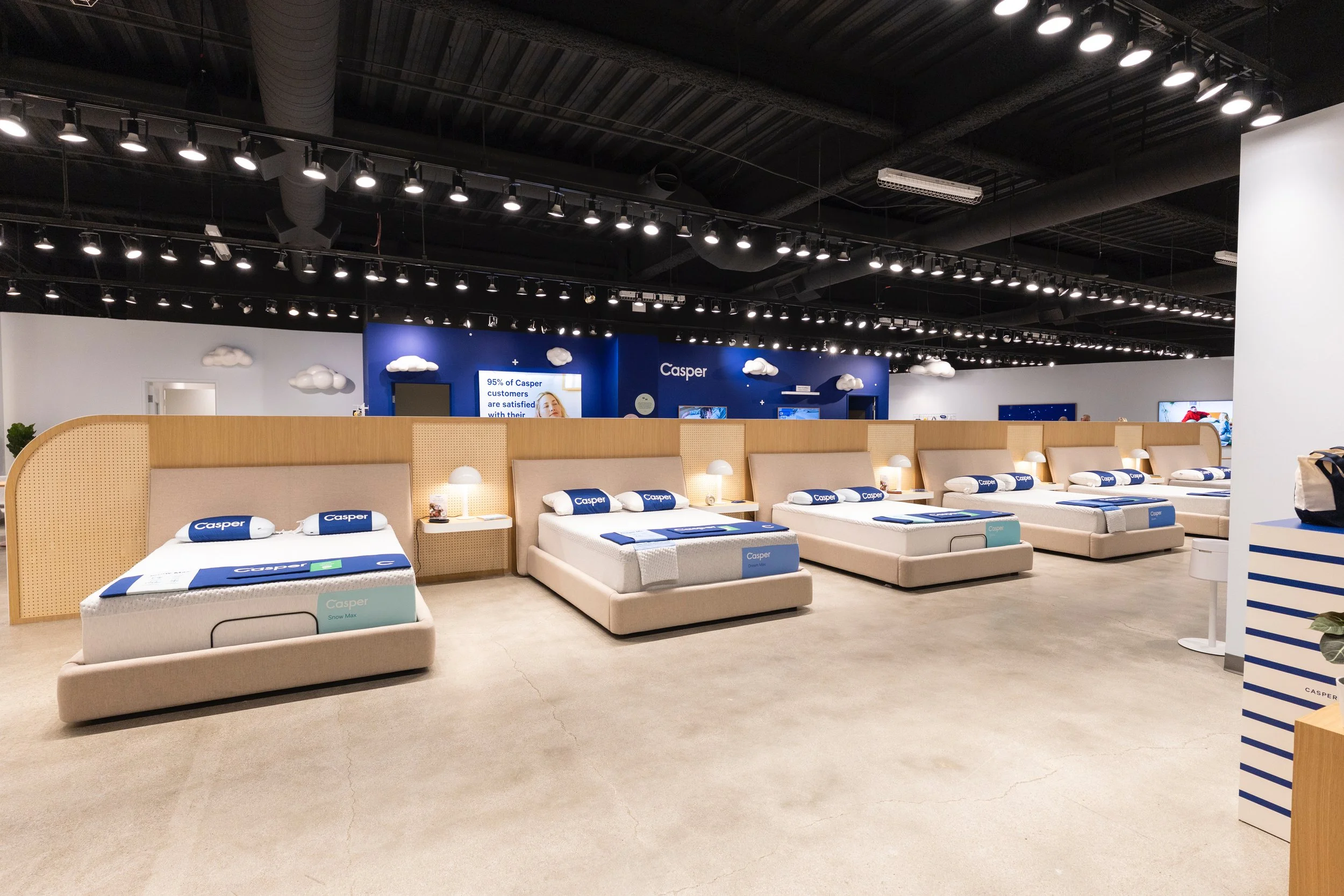 Showroom display of Casper mattresses arranged in rows, each with a Pillow and Bed frame, in a retail store with black ceiling and track lighting.