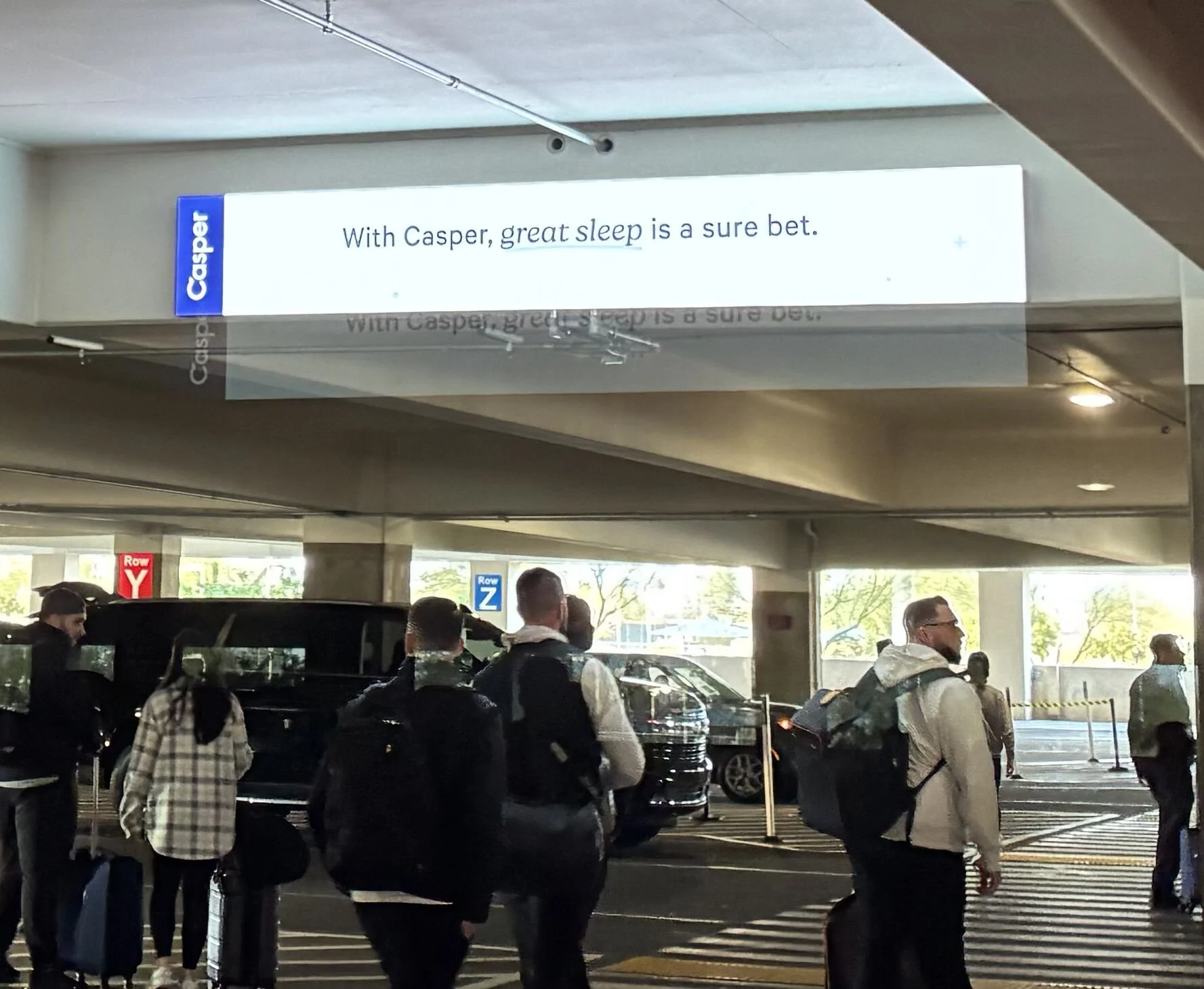 People walking in an airport terminal near parked cars, with a sign reading 'With Casper, great sleep is a sure bet.'