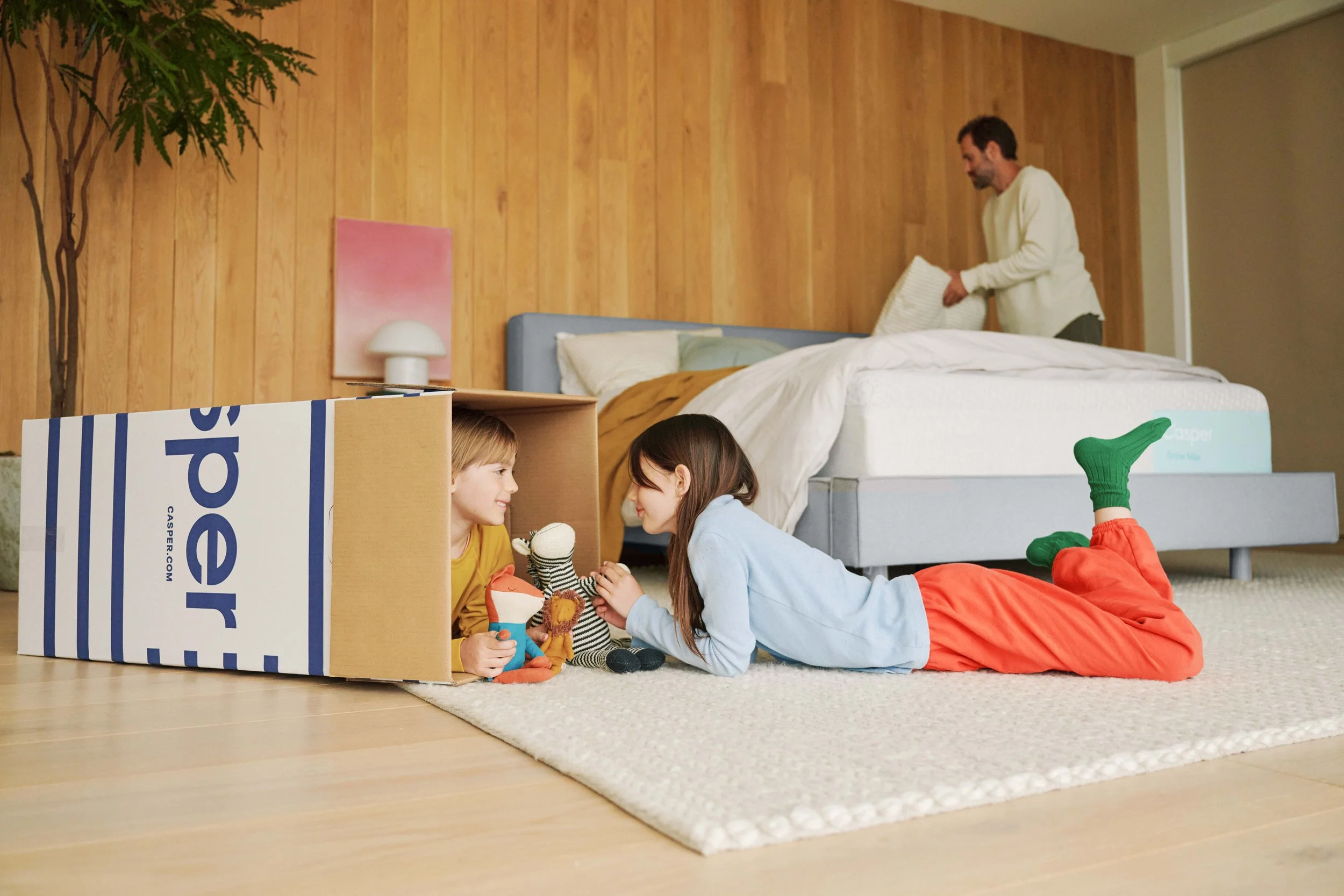 Two children playing with toys inside a cardboard box in a room with a wooden wall and a bed. One child is wearing an orange outfit with green socks, and the other is wearing a yellow shirt. An adult man is in the background making the bed.
