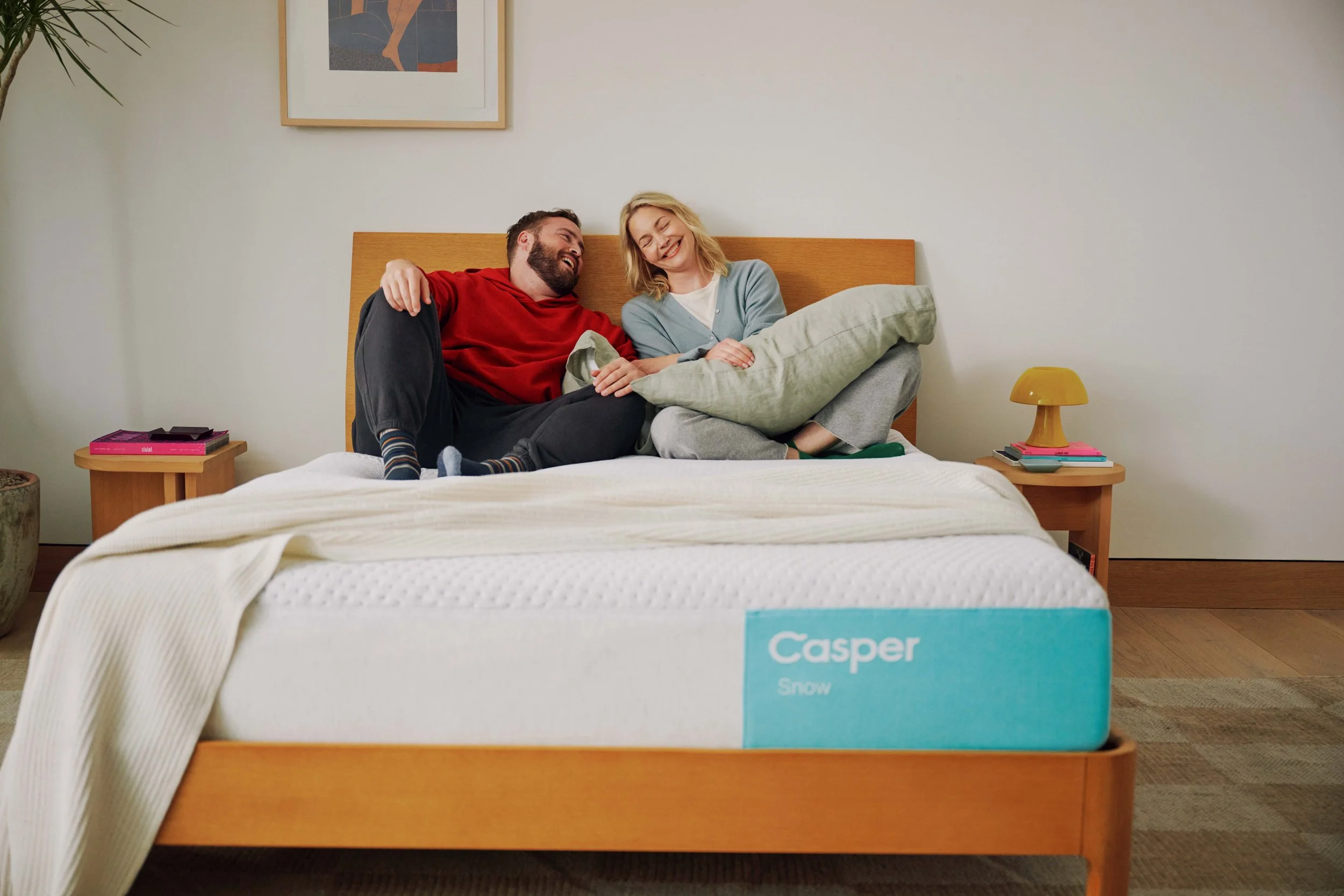 A man and woman sitting on a bed in a bedroom, laughing and hugging each other. The bed has a wooden headboard, and a pillow is held by the woman. There is a nightstand with a yellow lamp, books, and a remote control to the right of the bed. A potted plant is partially visible on the left, and a framed abstract artwork hangs on the wall above the bed.