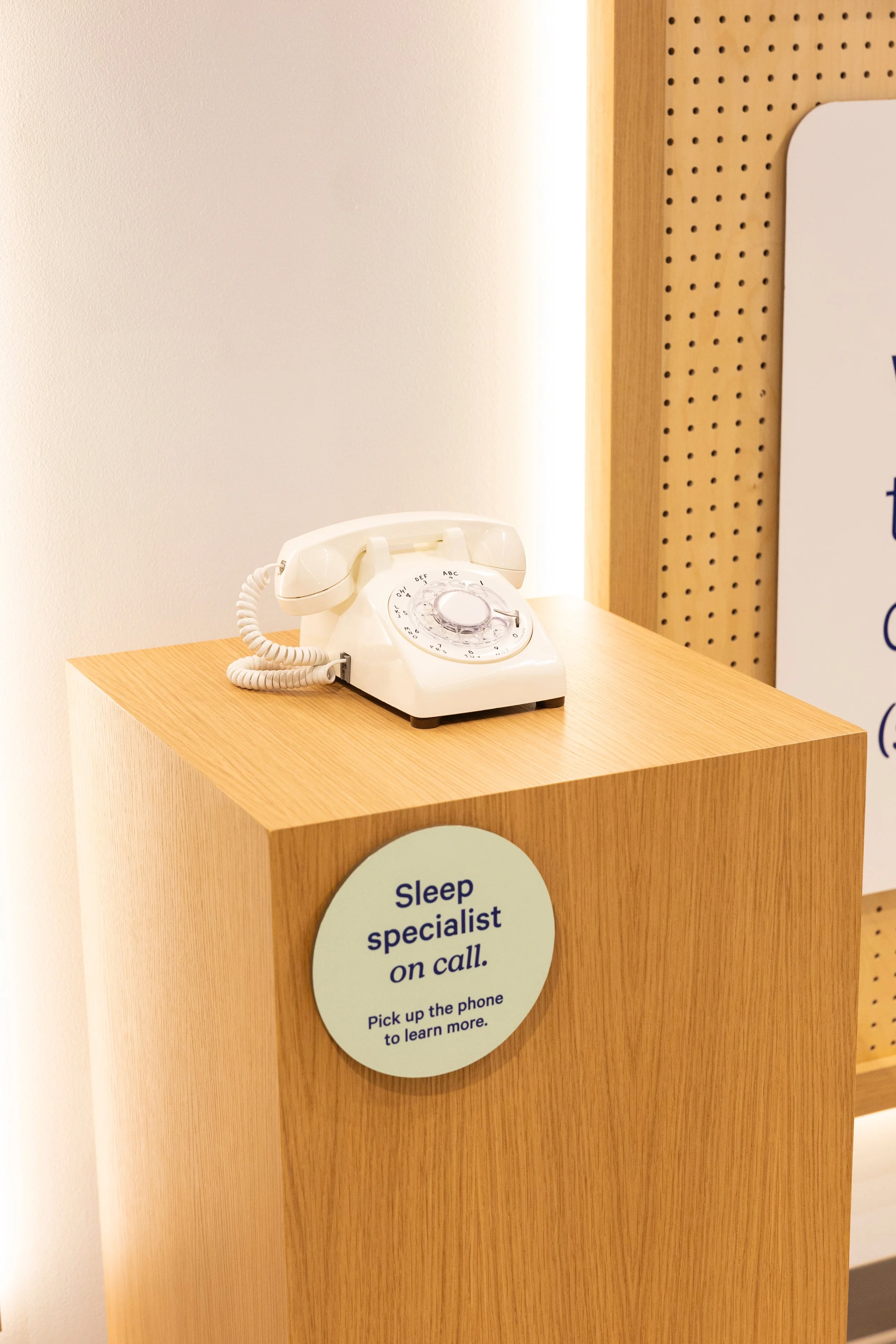 A white rotary dial telephone placed on a light wood table. The background has a white wall and a perforated wood panel with a sign that says 'sleep specialist on call.'