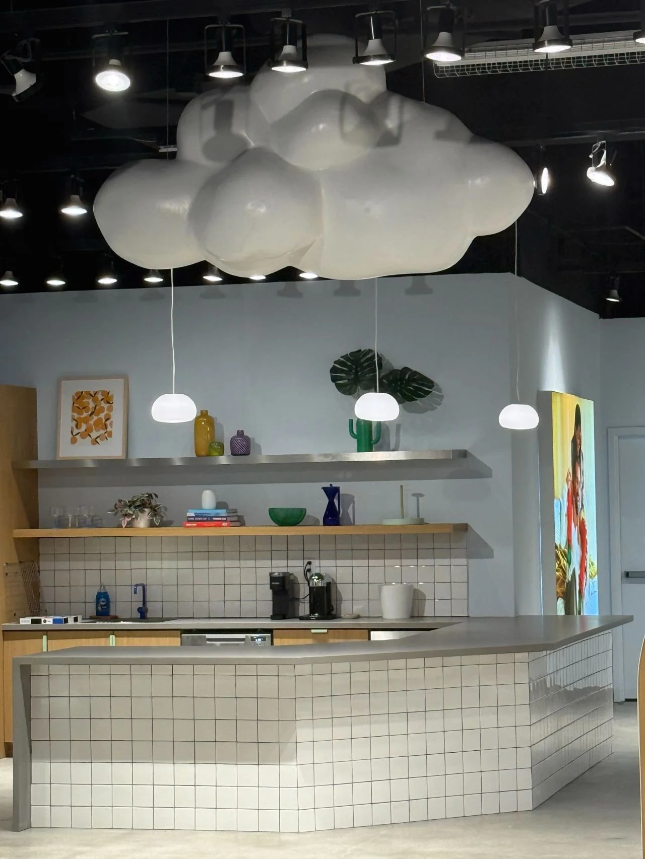 Interior of a modern cafe or kitchen with a cloud-shaped ceiling light, pendant lights, open shelves with decorative items, a coffee station, and a tiled counter.