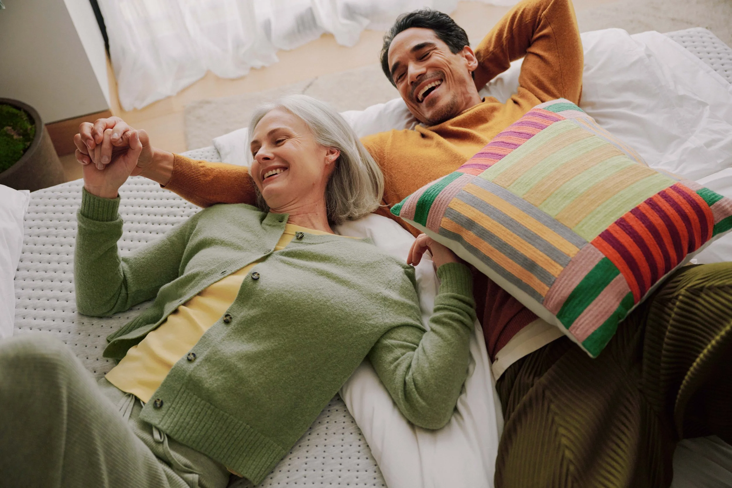 A happy older woman and a younger man lying in bed together, sharing a tender moment and holding hands, with both smiling.