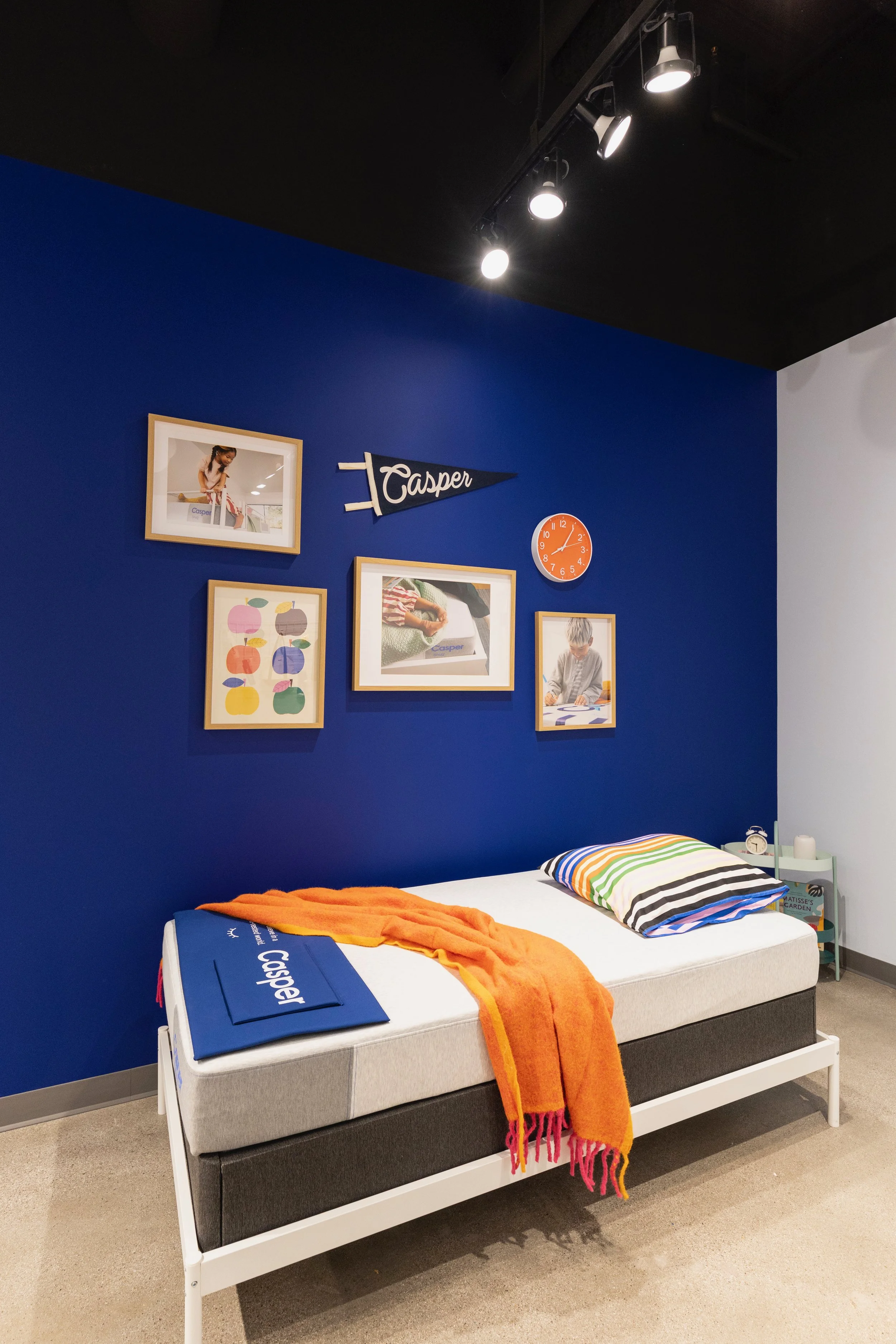 A retail store display featuring a mattress with striped pillow and orange blanket, blue wall background with framed photos, a small side table with books and a clock, and ceiling spotlights.