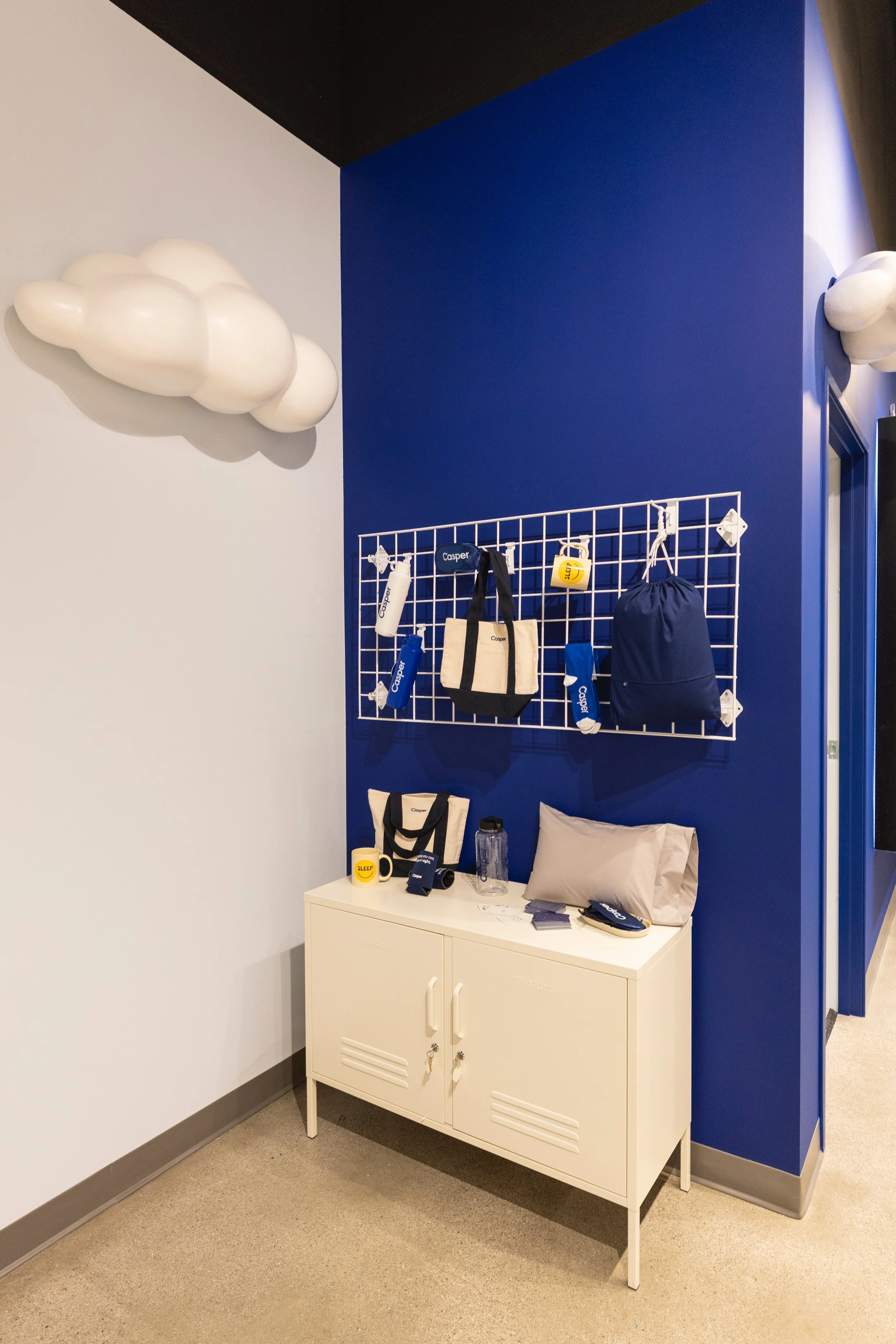 A corner of a room with a white cabinet, a blue wall, and a white grid wall organizer holding various items. There are cloud-shaped light fixtures on the walls, and a table with a mug, water bottle, and miscellaneous objects.