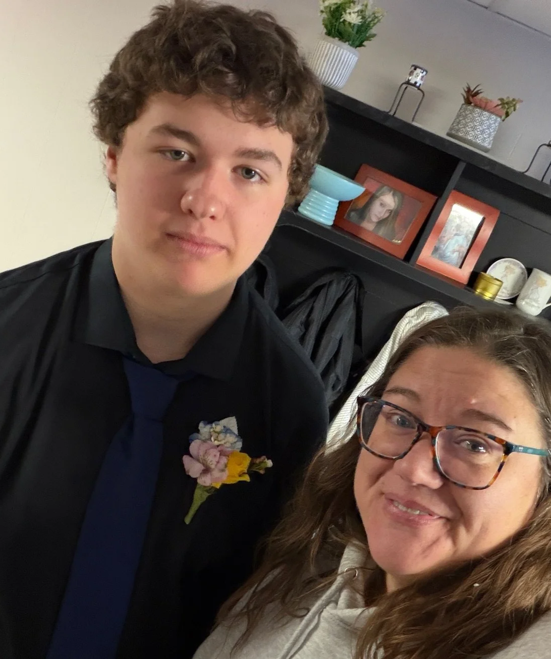 A young man with curly hair wearing a black shirt and a blue tie, and a woman with glasses and long hair, smiling for a selfie indoors.