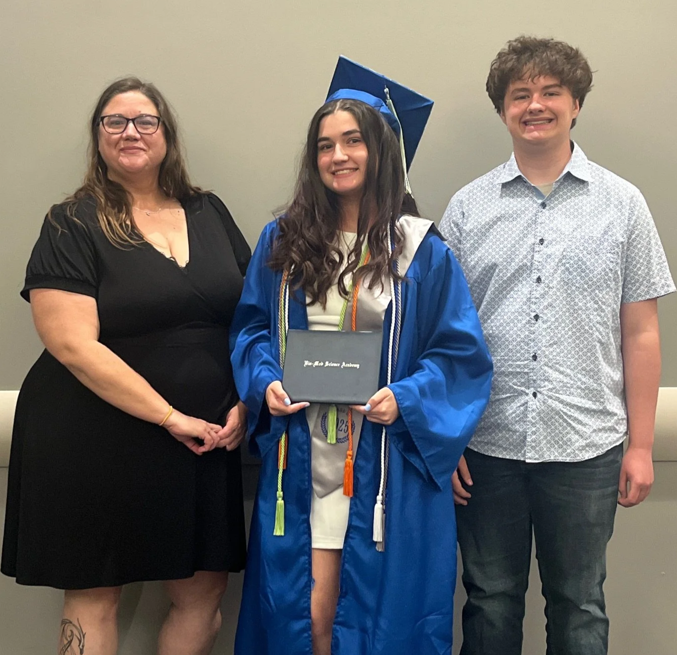 A young woman in a blue graduation gown and cap holding a diploma, standing between a woman in a black dress and a young man in a white patterned shirt, all smiling