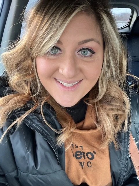 Close-up of a smiling blonde woman with blue eyes, wearing a black jacket and a brown shirt, sitting in a vehicle.