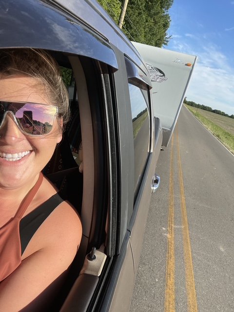 Woman with sunglasses smiling, leaning out of a side window of a pickup truck, on a rural road with truck's trailer in the background.