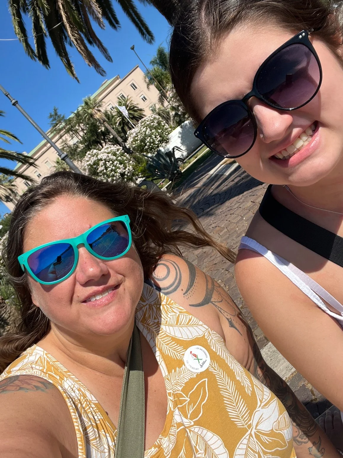 Two women wearing sunglasses taking a selfie outdoors, with a sunny sky, trees, and a building in the background.