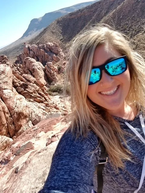 Woman with blonde hair wearing sunglasses and a navy jacket, smiling, in a rocky desert landscape with mountains in the background.