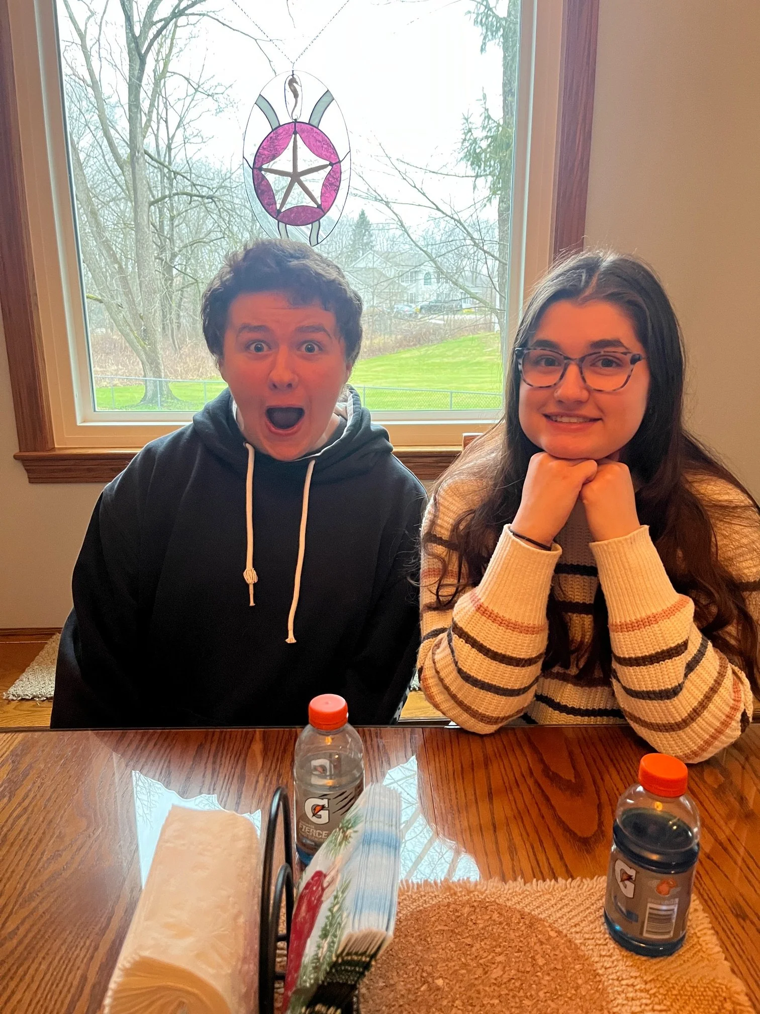 A young man and woman sitting at a wooden table inside a house, with a large window behind them showing leafless trees and grass outside. The man has short dark hair, is wearing a black hoodie, and has an excited expression with his mouth open. The woman has long dark hair, wears glasses, a beige and brown striped sweater, and is smiling with her chin resting on her hands. There are two Gatorade bottles, a roll of paper towels, and a box of tissues on the table.