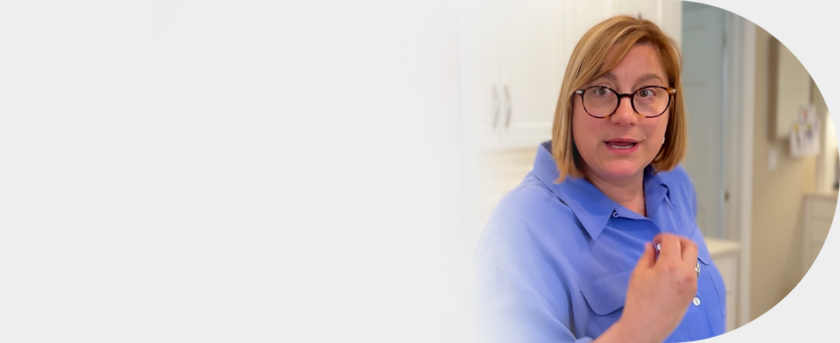 A woman with shoulder-length blonde hair, glasses, and a blue shirt making a fist gesture and speaking in a kitchen setting.