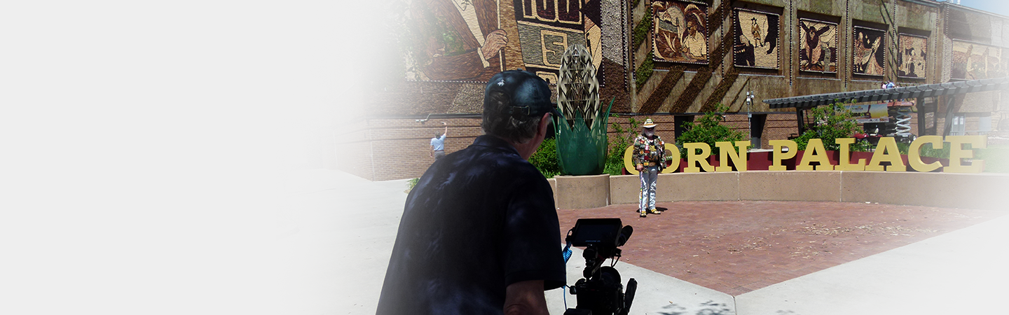 Man operating a camera outdoors near a large sign that reads 'CORN PALACE,' with artwork and murals on the building behind him.